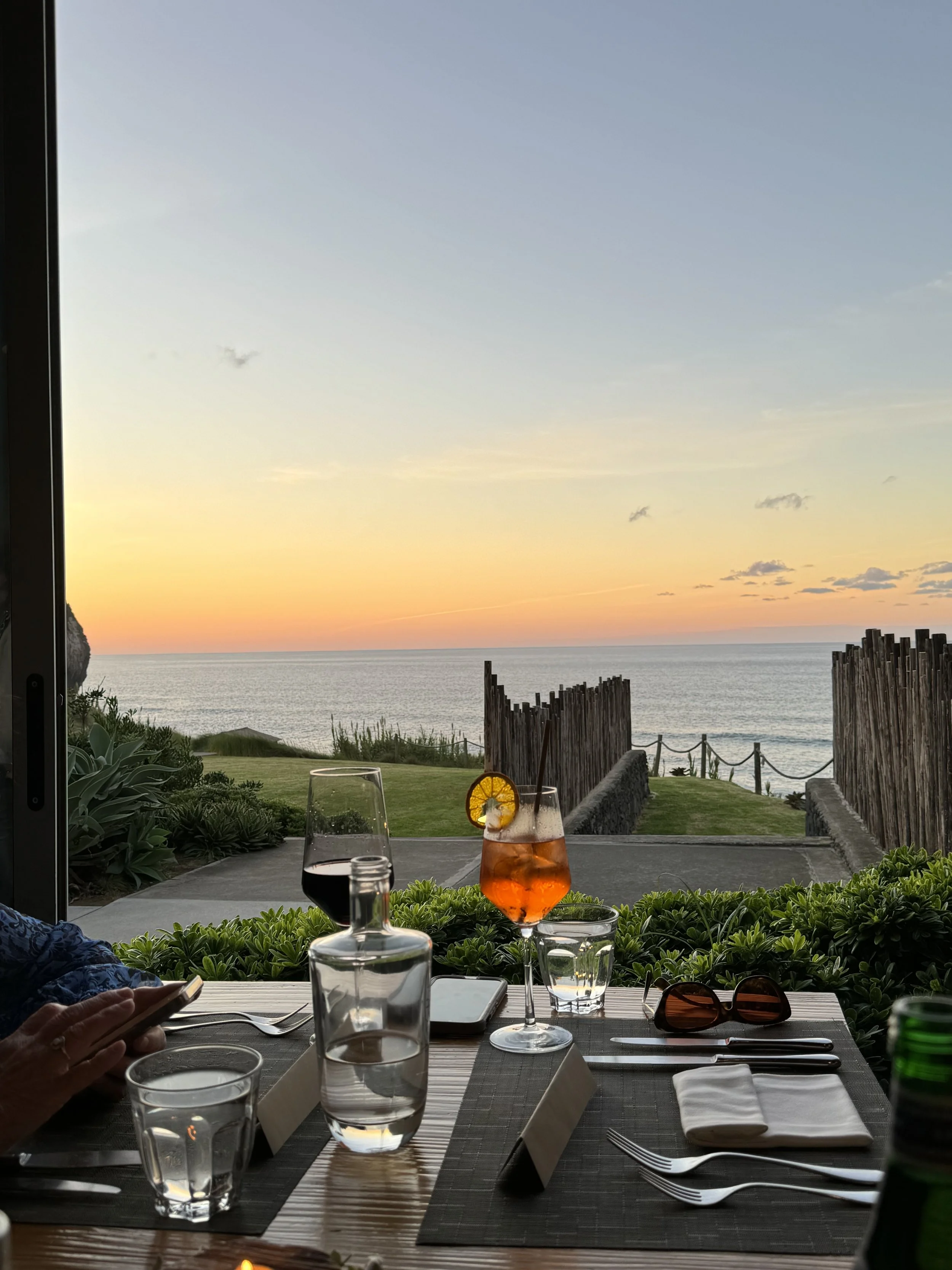 A table set for dining with glasses of wine and drinks, overlooking a scenic view of the ocean during sunset, with a partly cloudy sky and seaside landscape in the background.