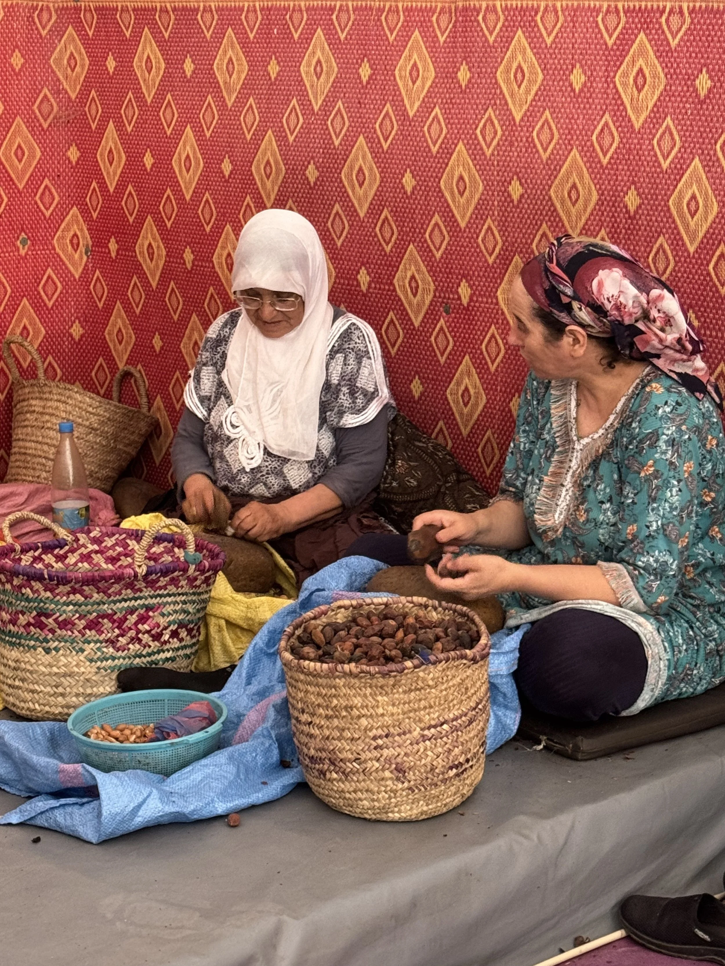 Two women sitting on the ground at a market stall, selling nuts and dried fruits in colorful baskets. One woman is wearing a white headscarf and glasses, and the other is wearing a headscarf with pink flowers. They are surrounded by baskets filled wi