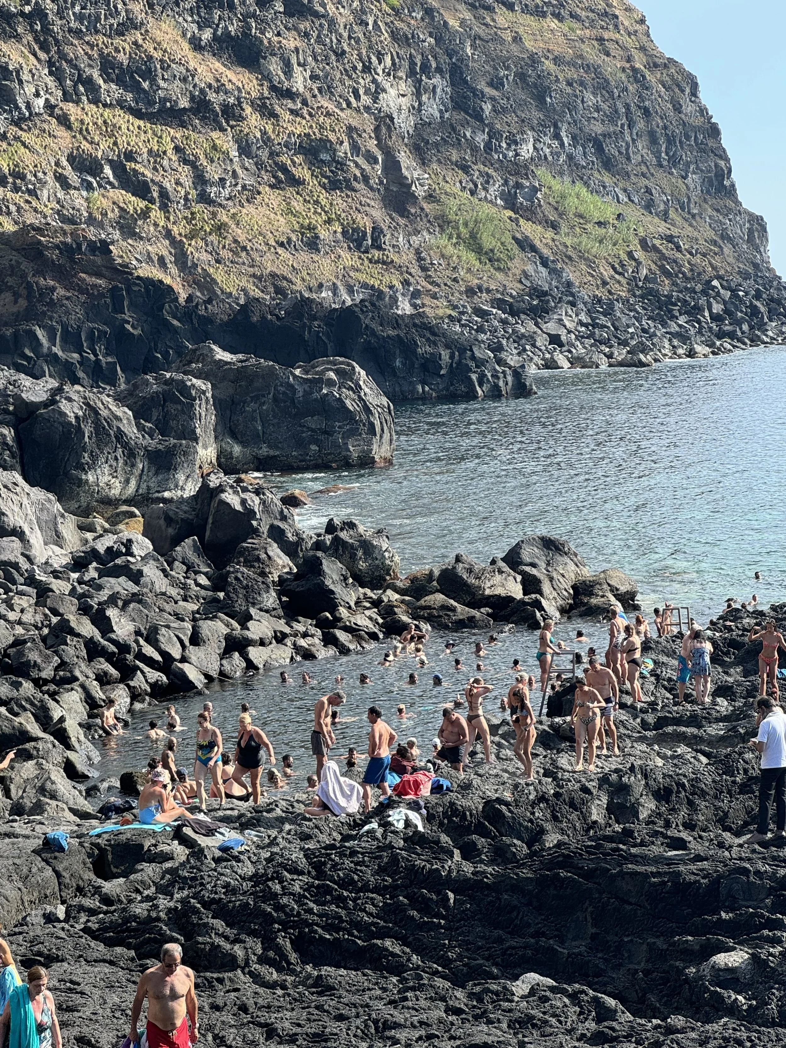 A rocky shoreline with many people swimming and relaxing in the water and on the rocks, surrounded by steep cliffs and clear skies.
