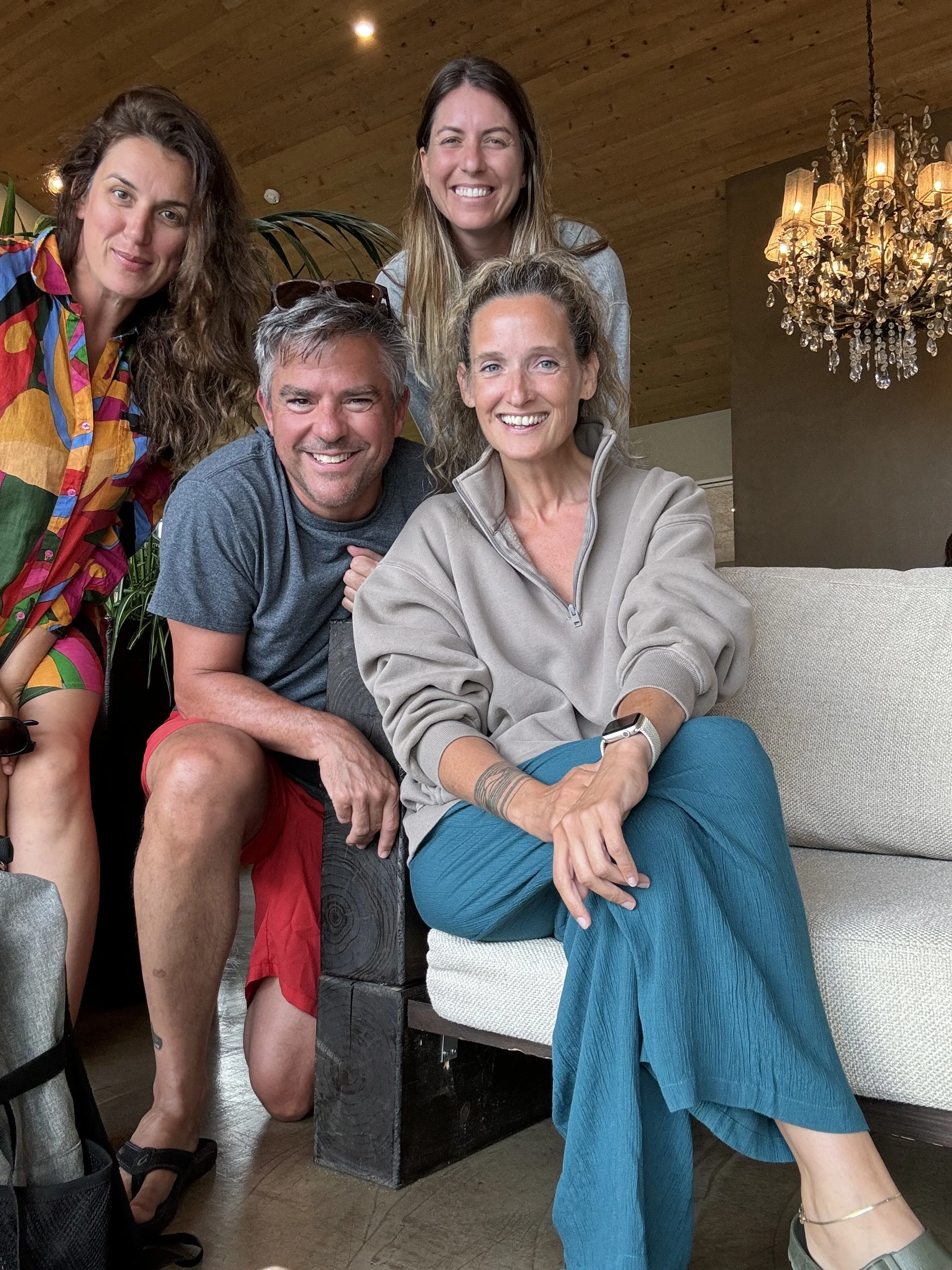 Group of five happy adults in a cozy indoor setting, with wooden ceiling and chandelier, sitting and standing close together, smiling at the camera.