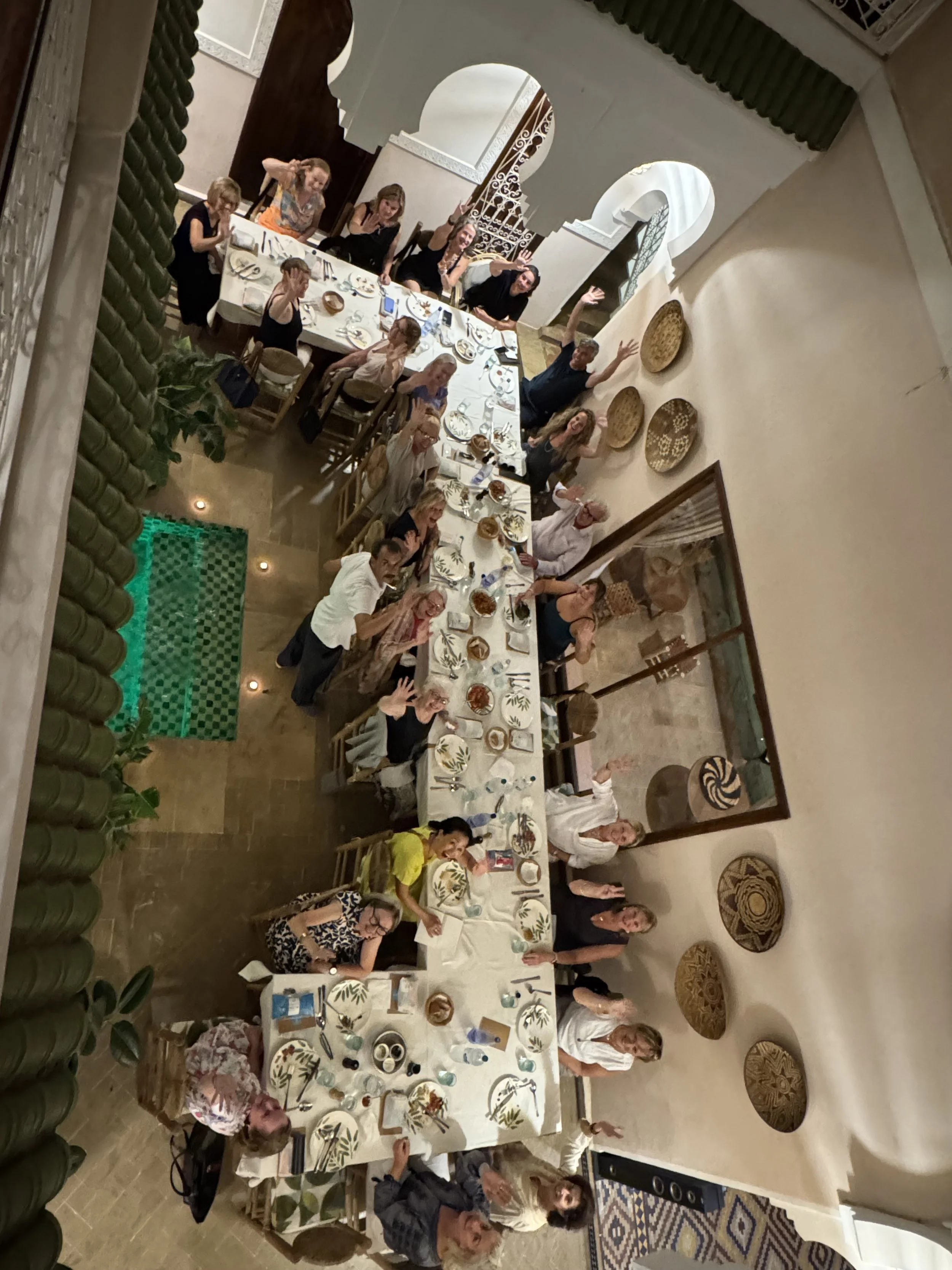 People gathered around a long dining table in a restaurant, waving and smiling at the camera, with decorative wall art and green plants in the background.