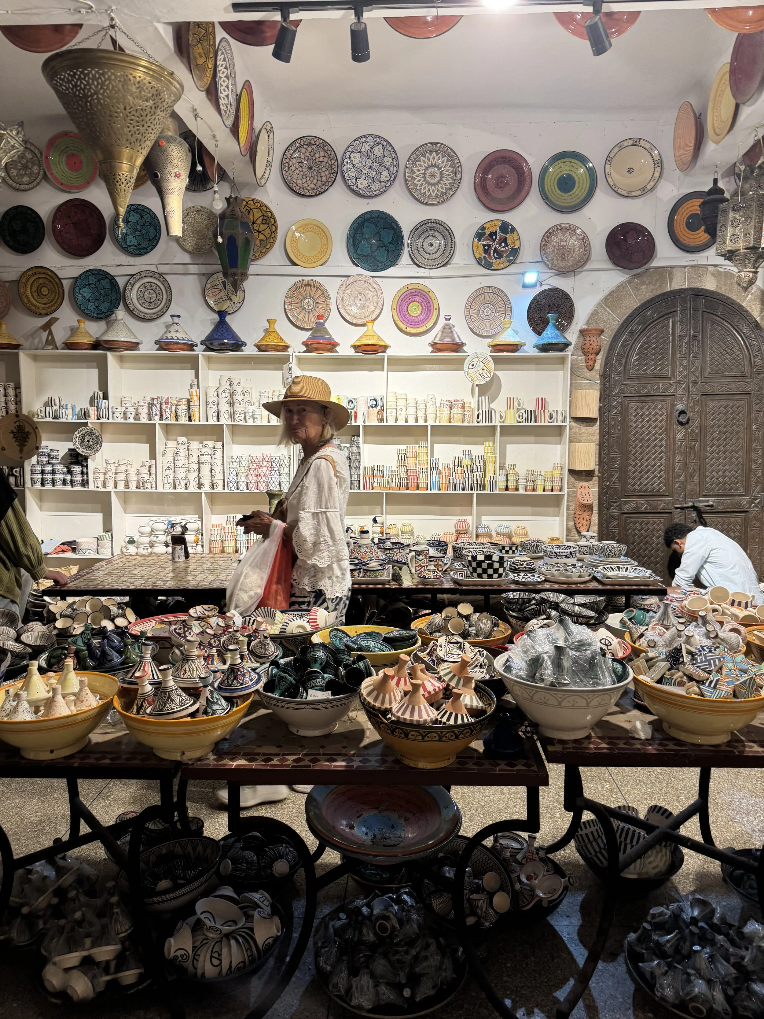 A woman with a straw hat looks at a display of patterned ceramics, including bowls, cups, and decorative plates, in a shop with shelves and a large ornate wooden door in the background.
