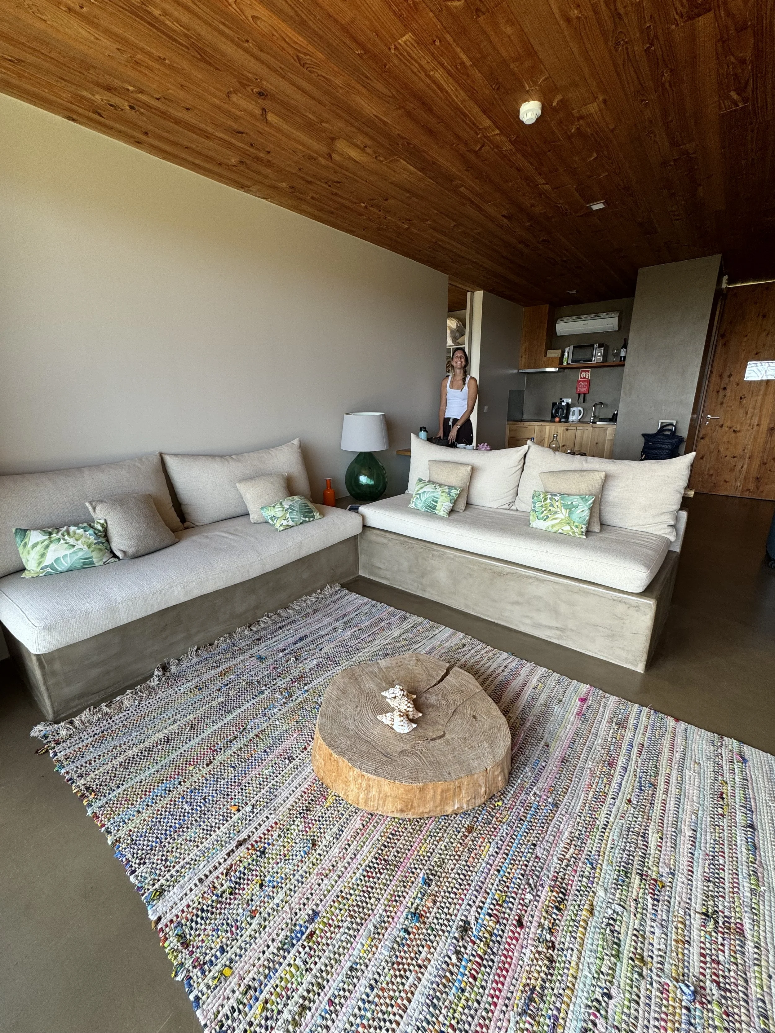 Living room with beige sectional sofa decorated with green and beige pillows, a colorful woven rug, and a round wooden coffee table with seashells on top. In the background, a woman in a white top stands near a kitchen area with wooden cabinets, appl