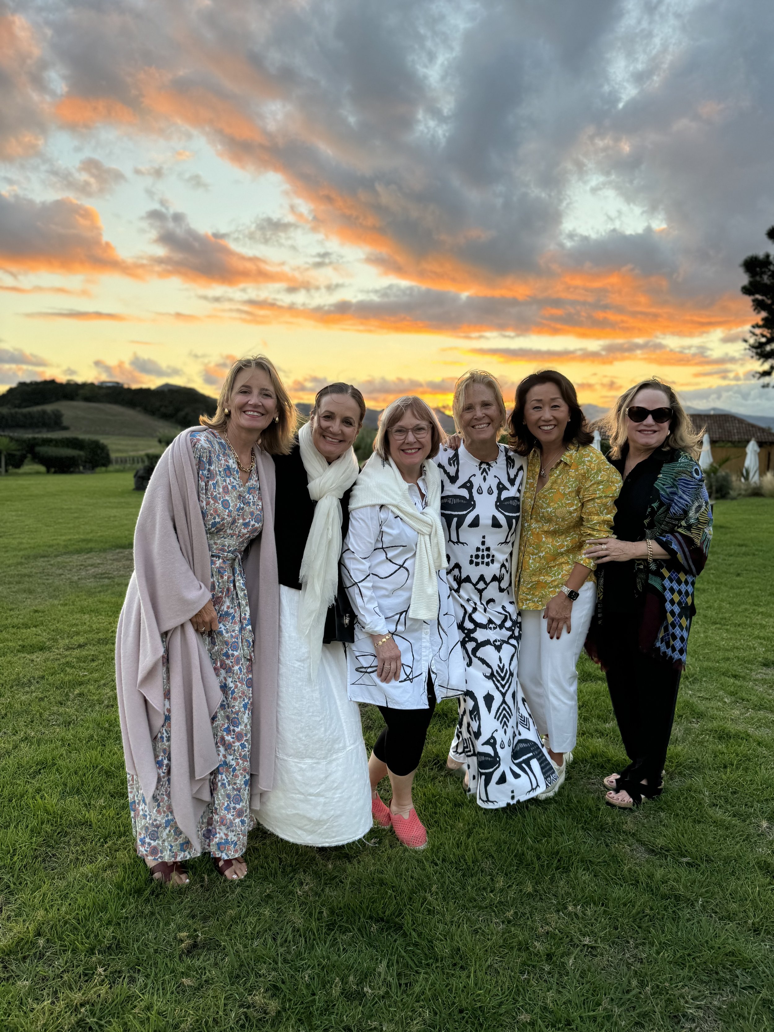 Six women standing on grass with sunset sky in the background.