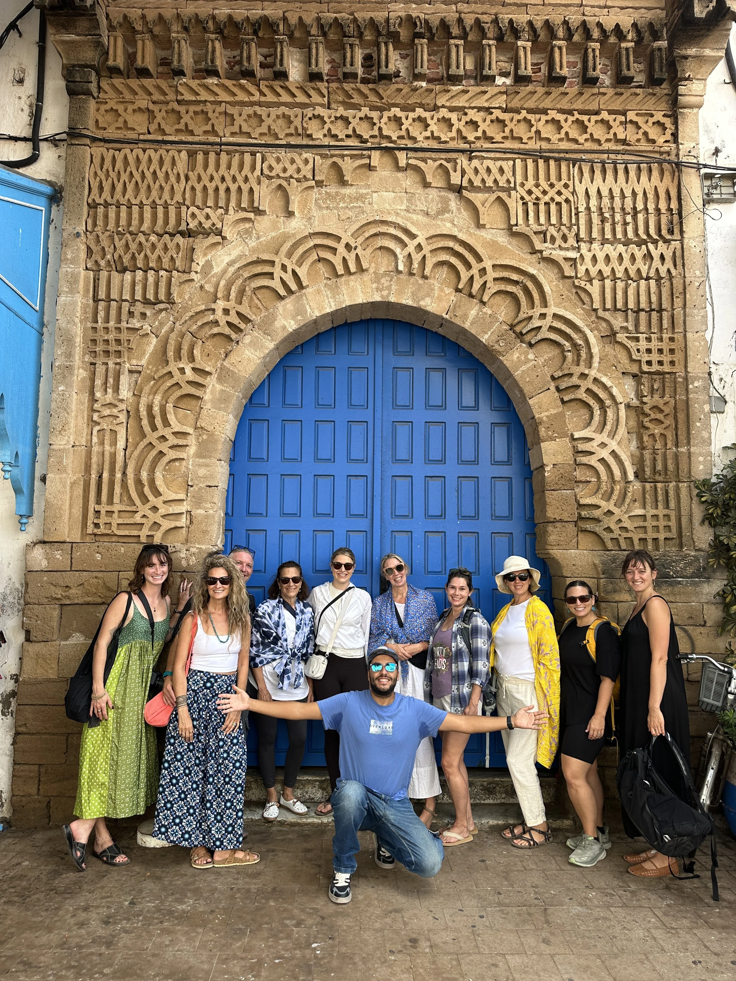A group of twelve diverse people, including men and women of varying ages, posing happily in front of a large blue door with elaborate stone carvings above it. The group appears to be tourists or friends enjoying a sightseeing moment together.