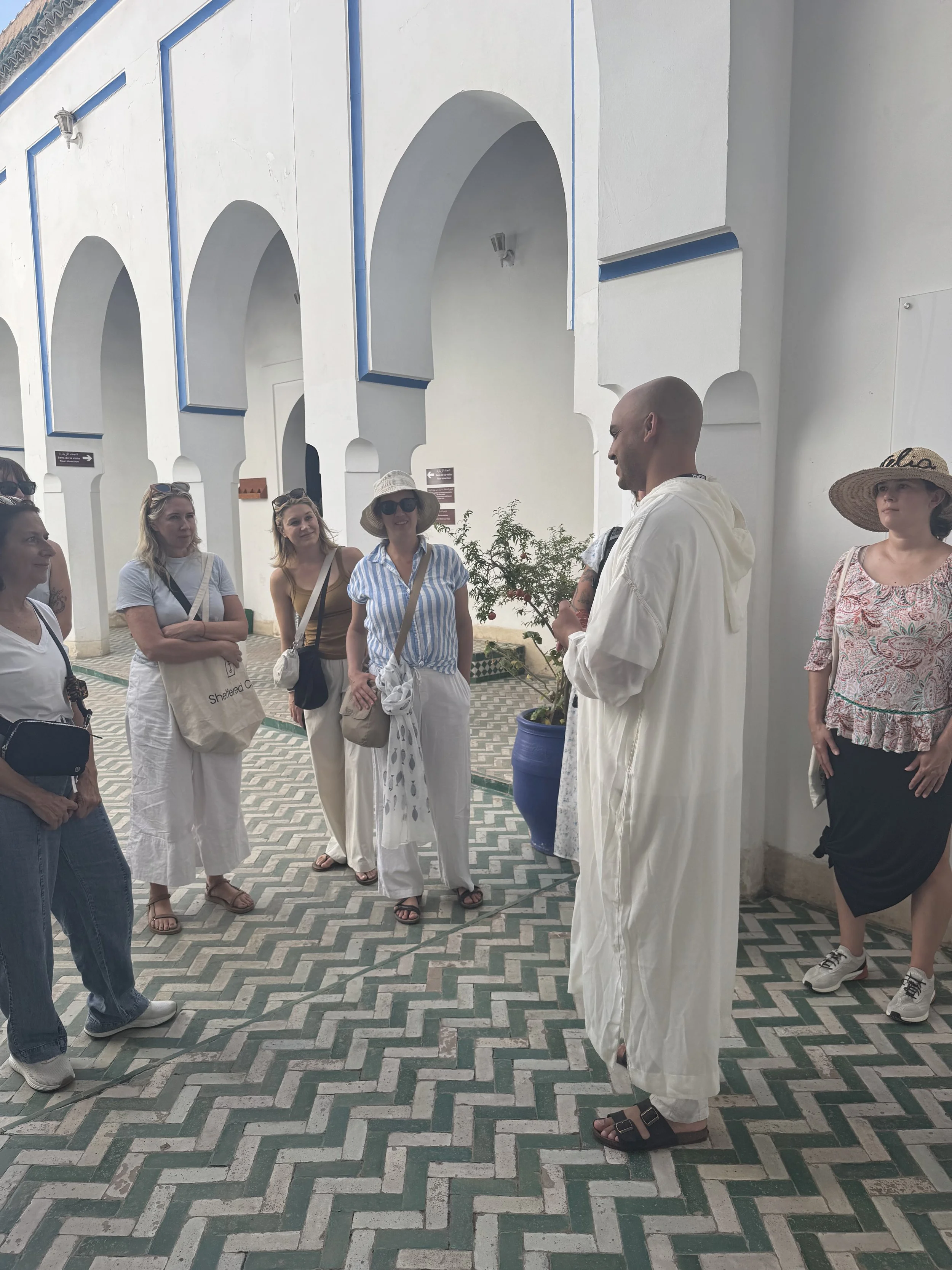 Group of people standing and listening to a guide in an outdoor area with white walls, arches, and a patterned tile floor.