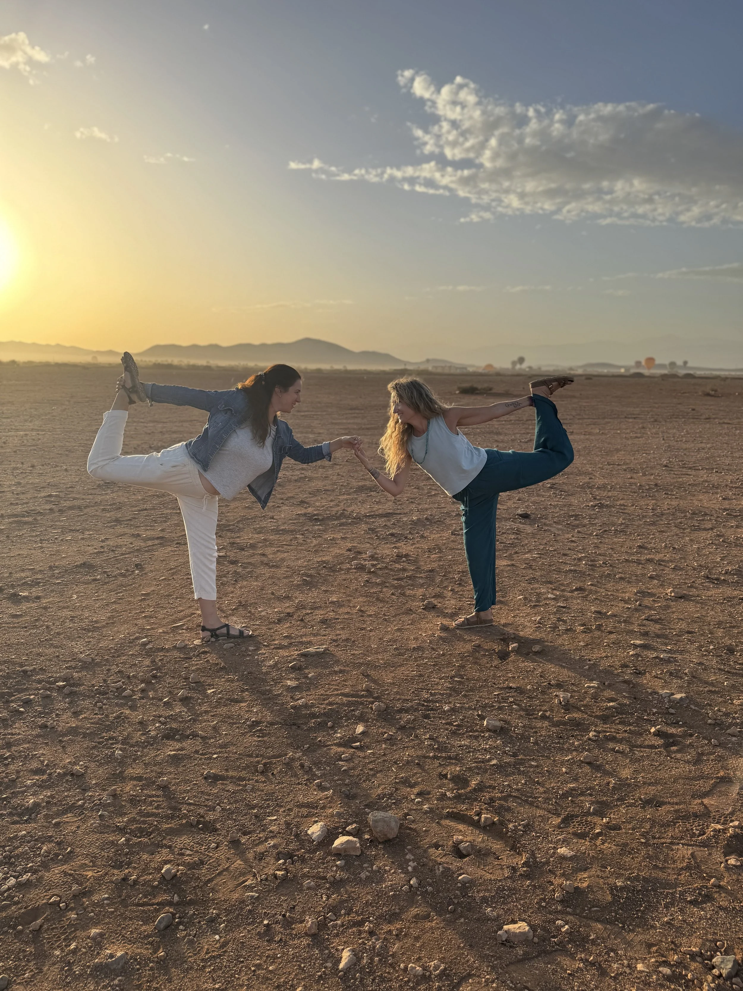Two women practicing yoga outdoors on a dirt field at sunset, holding each other's hands while doing balancing poses with one leg lifted behind them.