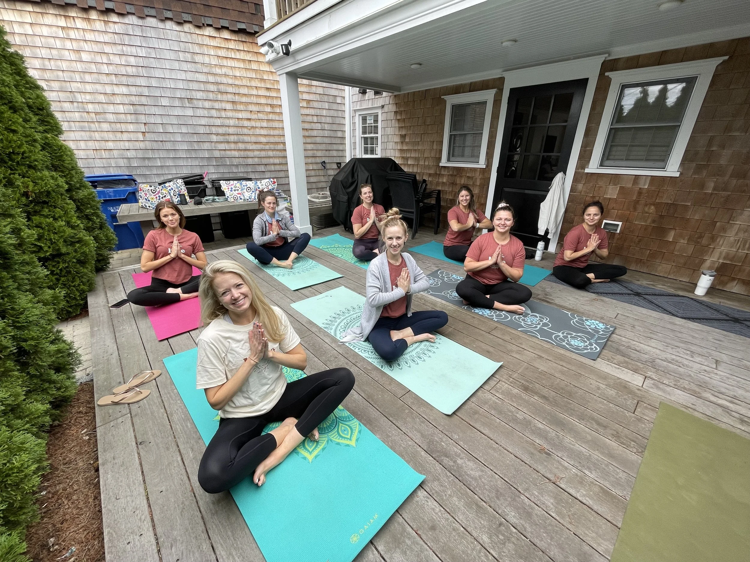 A group of people practicing yoga on a wooden deck in a backyard. They are sitting cross-legged with hands in prayer position, smiling, and surrounded by yoga mats.