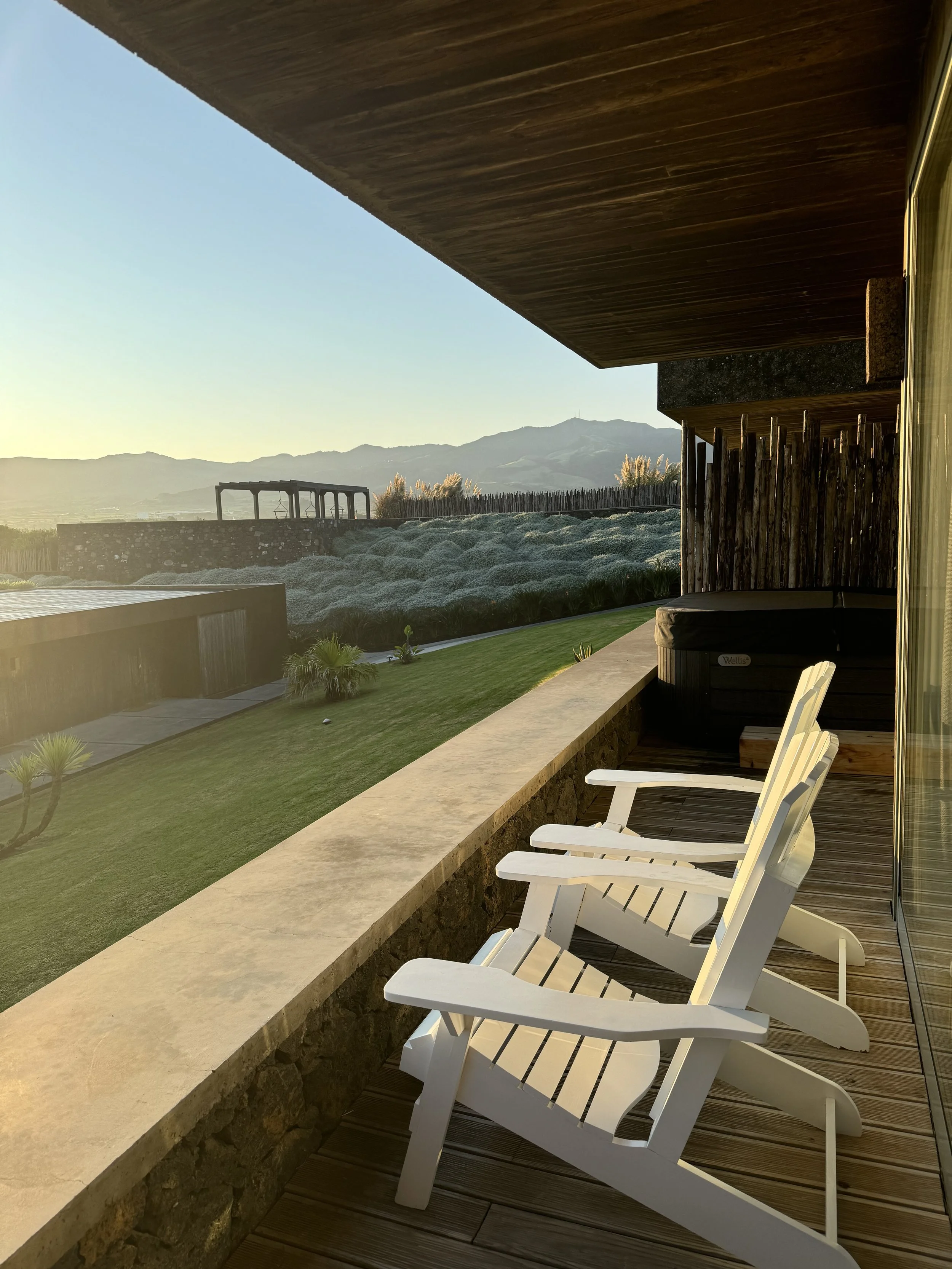 Four white outdoor lounge chairs on a wooden deck overlooking a grassy yard and distant mountains at sunset.