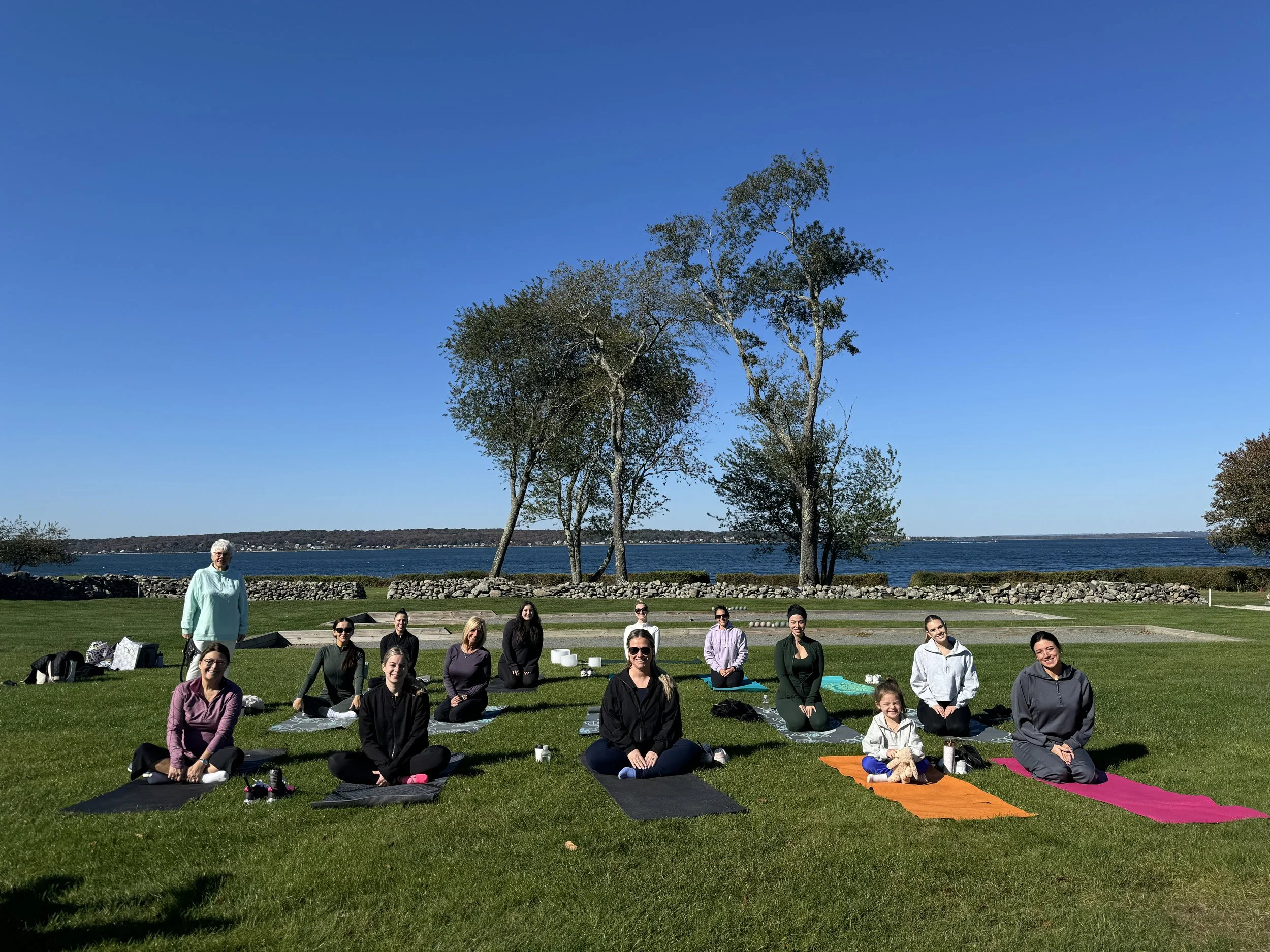 A group of people practicing yoga outdoors on a grassy area by a body of water, with blue skies and trees in the background.