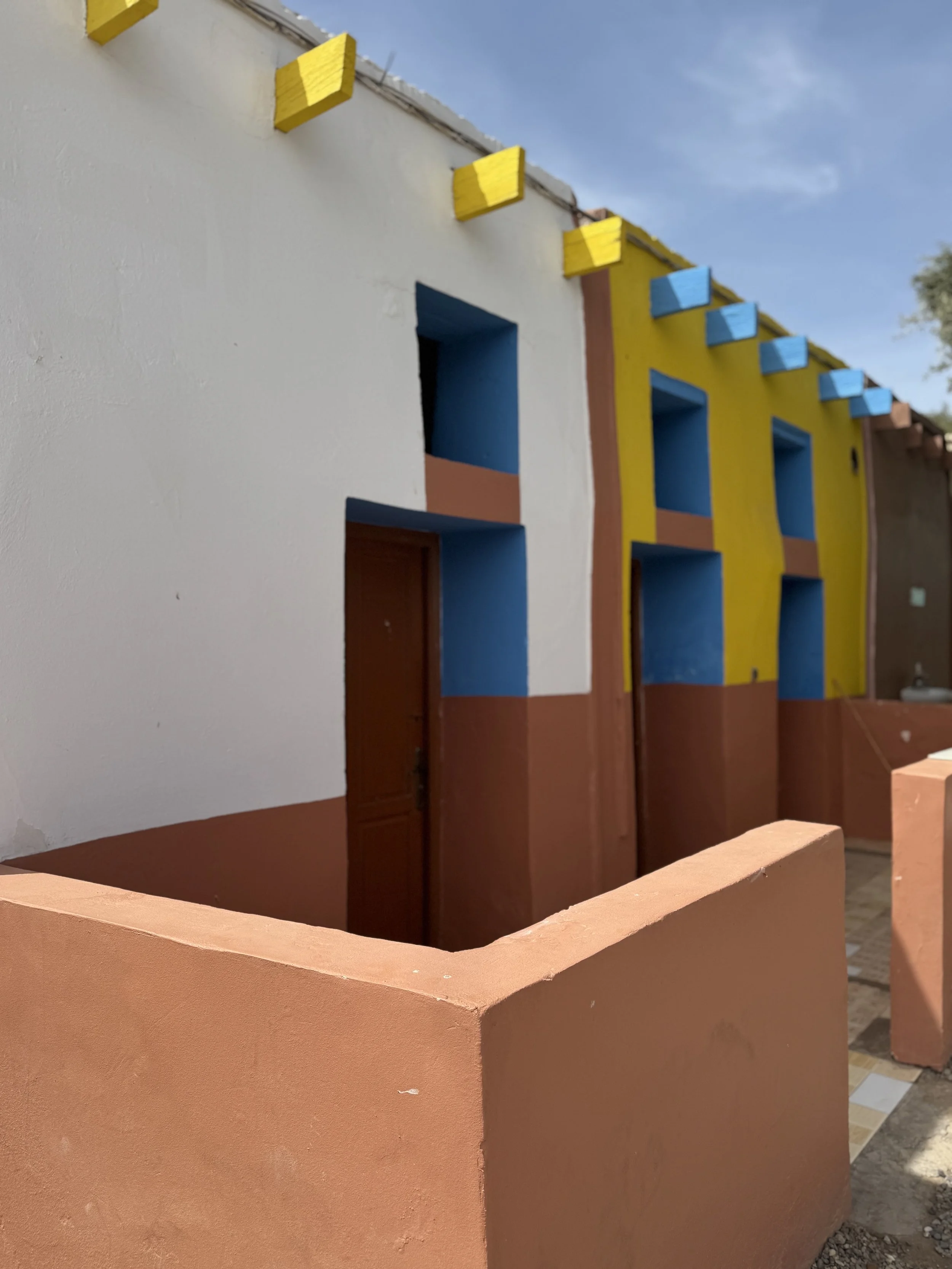 Colorful building with yellow, blue, and terracotta sections, featuring geometric windows and a curved terracotta-colored wall in the foreground under a blue sky.