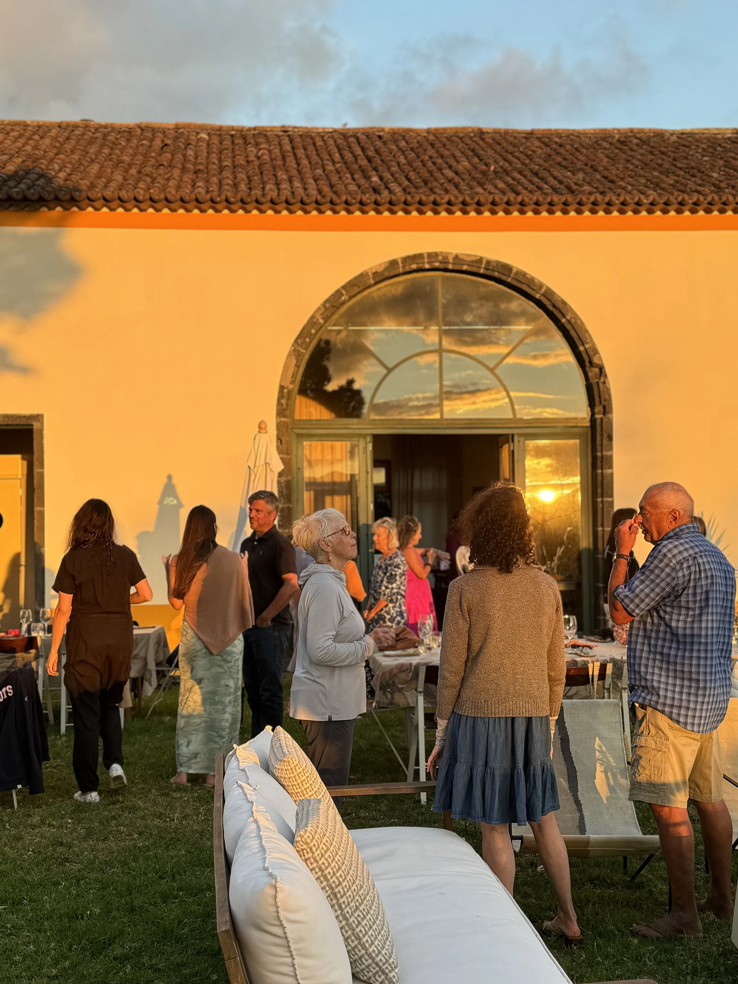 People gathered outdoors during sunset, socializing and enjoying a casual event with a building having a large arched window reflecting the sky and sunset.