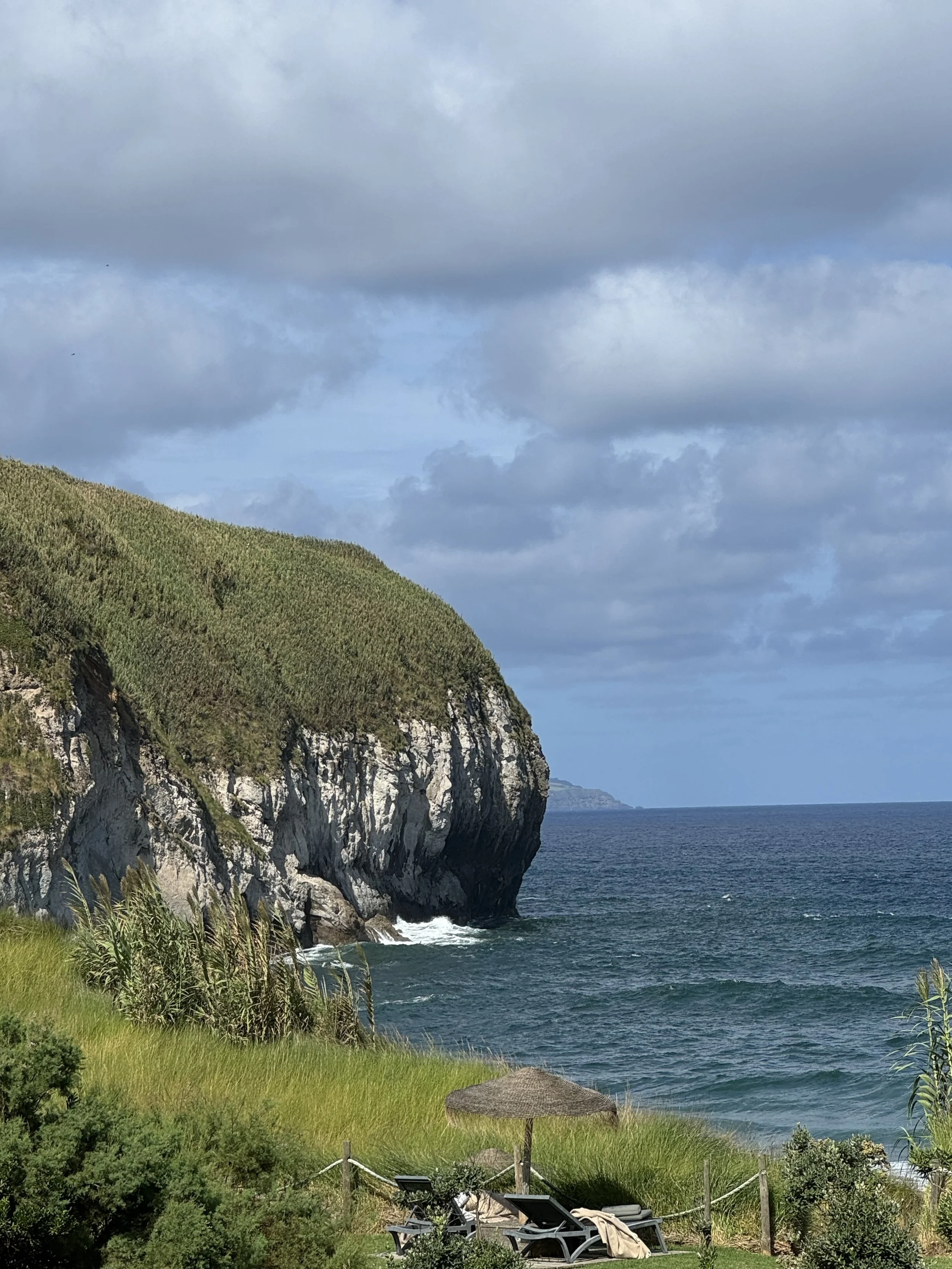 A scenic coastline with a grassy area in the foreground, a thatched umbrella, lounge chairs, a wooden fence, a rocky cliffside covered with greenery, and the ocean under cloudy skies.