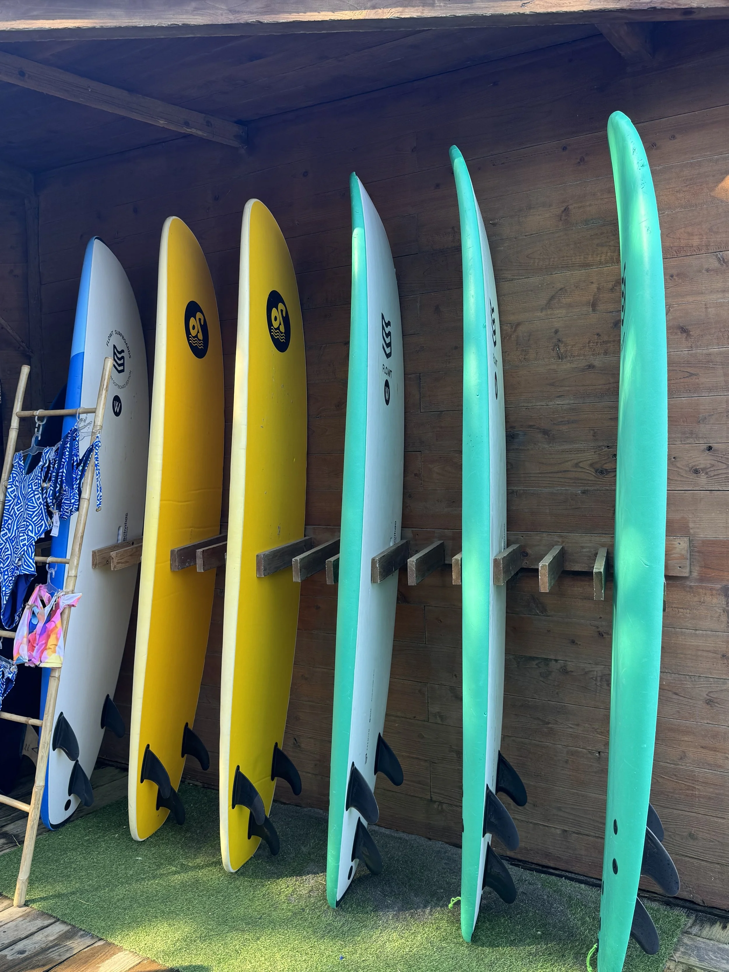 Surfboards in white, yellow, and turquoise are standing upright against a wooden wall, with a small rack of blue and pink swimwear to the left.