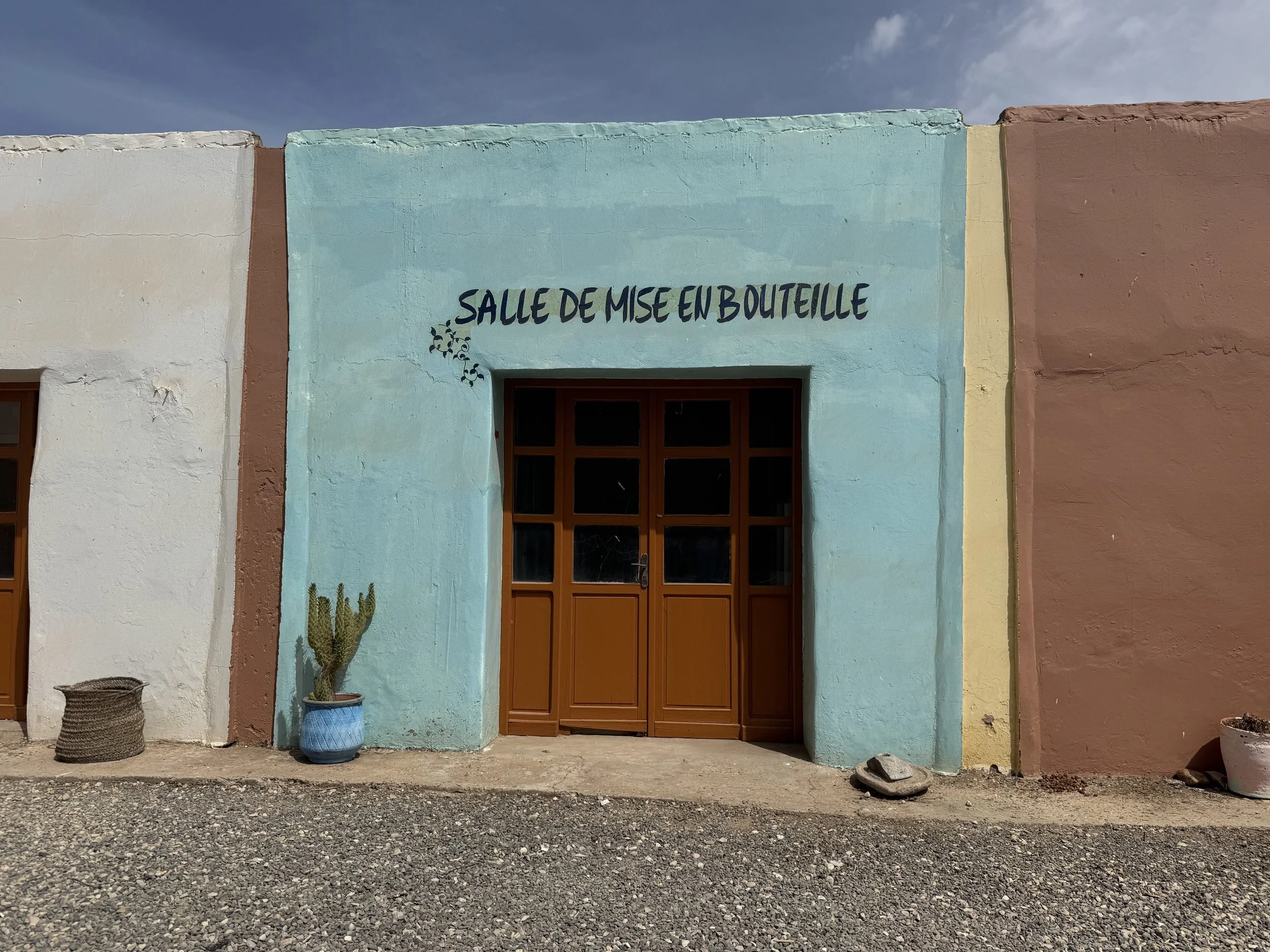 Front view of a colorful building with a sign in French reading 'Salle de mise en bouteille,' featuring a blue wall, a wooden door, potted plants, and baskets.