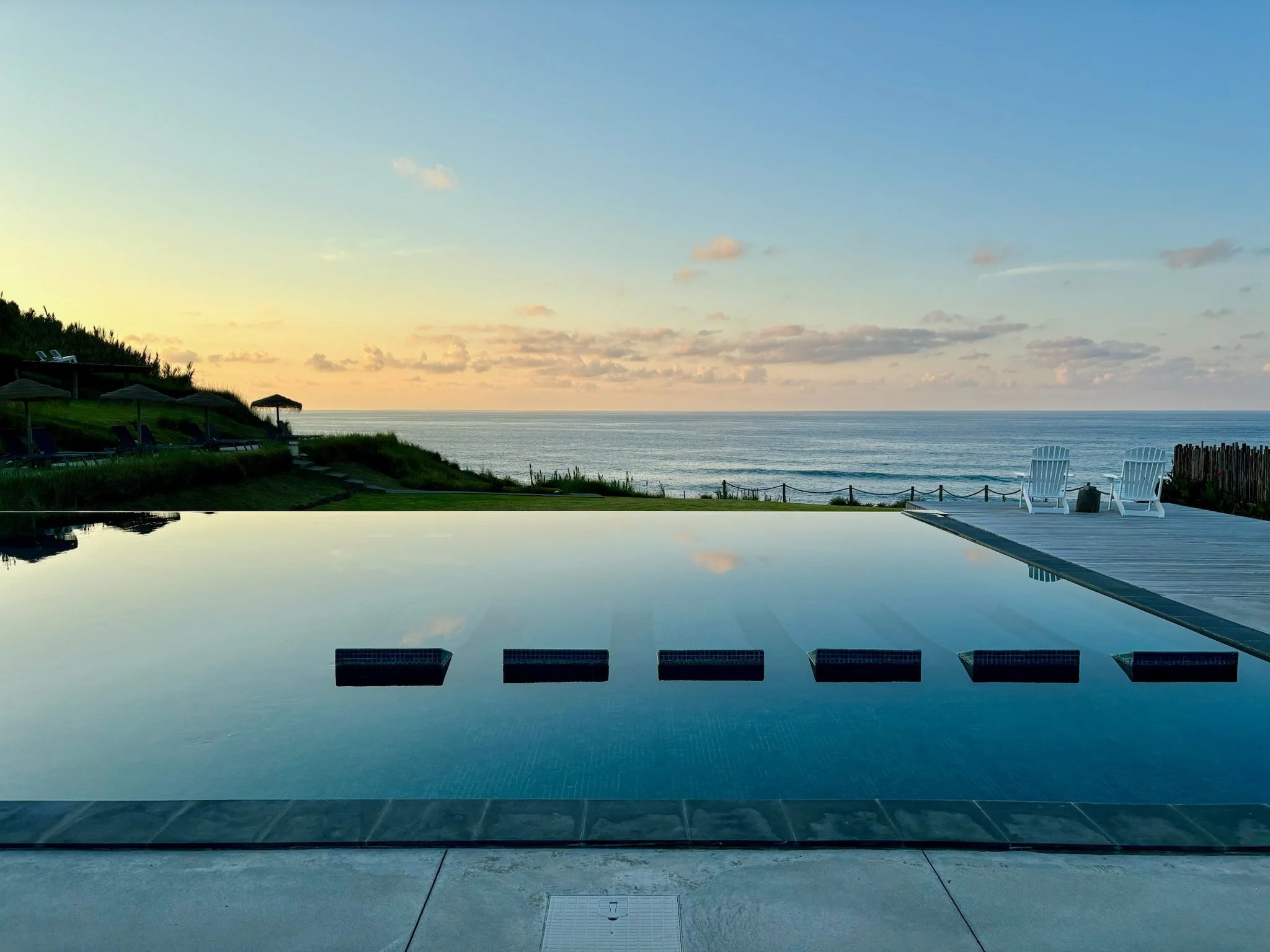 View of a pool with a counter at the edge and several empty white chairs facing the beach and ocean under a partly cloudy sky at sunset.