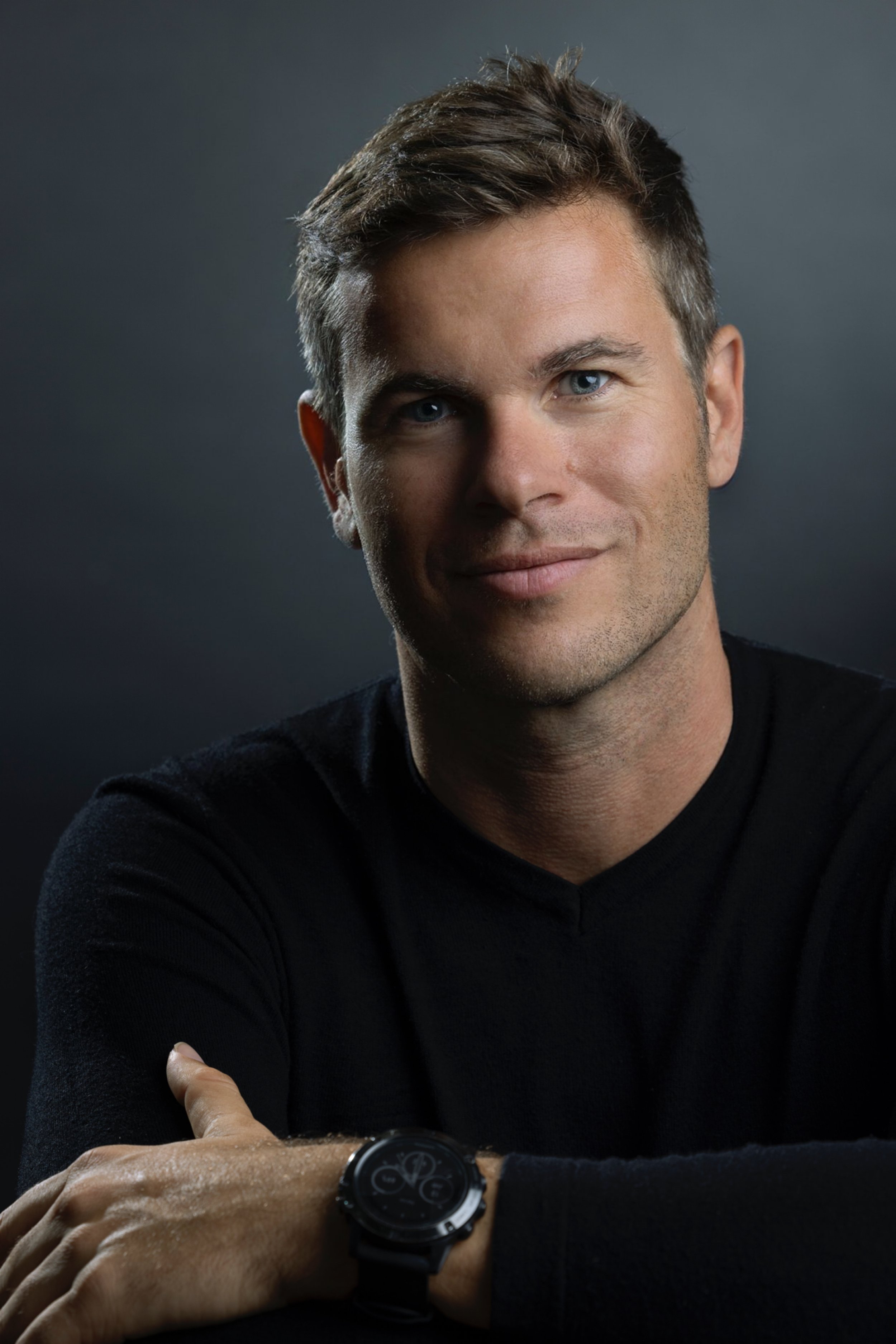 Hombre con cabello castaño, ojos azules, piel clara, vistiendo una camiseta negra, mostrando una expresión de confianza y sonriendo ligeramente, usando reloj inteligente en la muñeca izquierda, fondo oscuro.