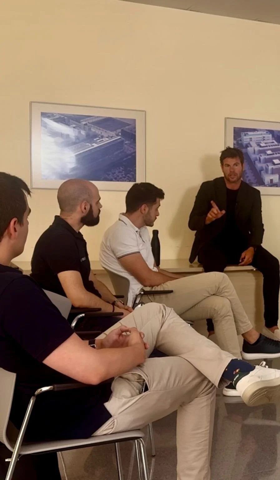 Un hombre de cabello oscuro y barba, sentado en la esquina, dando una charla a un grupo de jóvenes en una sala de reuniones. Cuatro personas en primer plano escuchando atentamente, con dos fotos de un complejo arquitectónico en la pared.