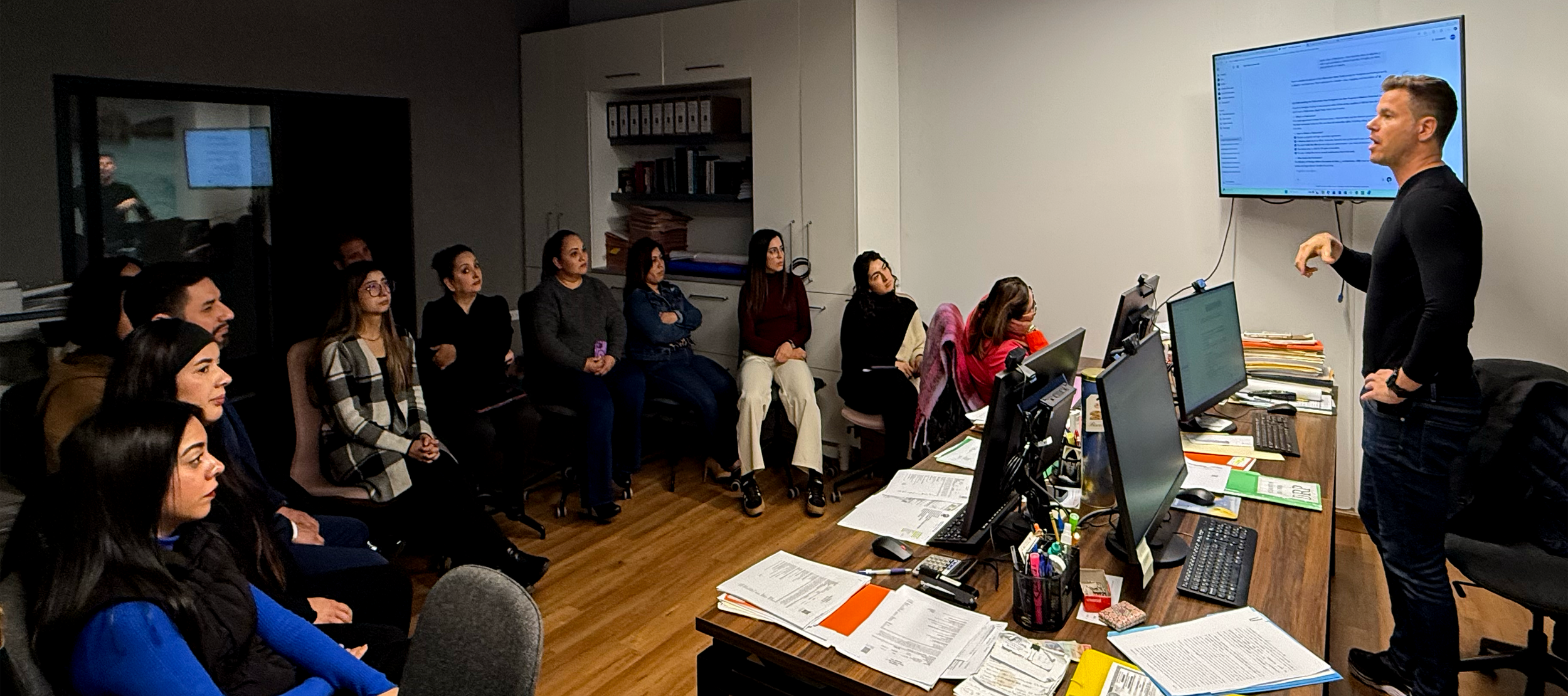 Un instructor dando una charla a un grupo de personas en una oficina con varias computadoras y documentos sobre la mesa.