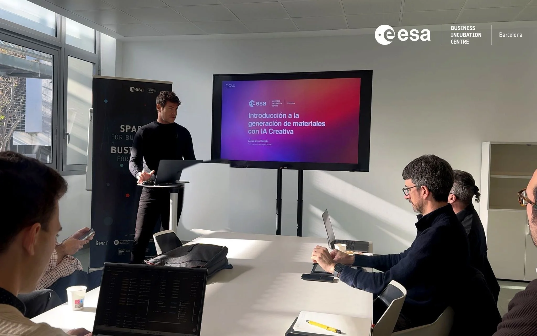 Presentación en un salón de reuniones con una pantalla grande que muestra el título 'Introducción a la generación de materiales con IA Creativa' durante un evento de incubación de negocios en Barcelona organizado por la Agencia Espacial Europea, con asistentes atentos y tomando notas.