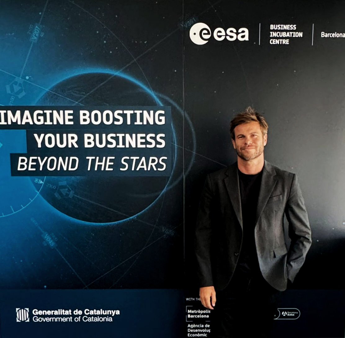 Hombre sonriente con chaqueta negra de pie junto a cartel de la Agencia Espacial Europea en Barcelona, que promueve la incubación de negocios y el impulso empresarial más allá de las estrellas.