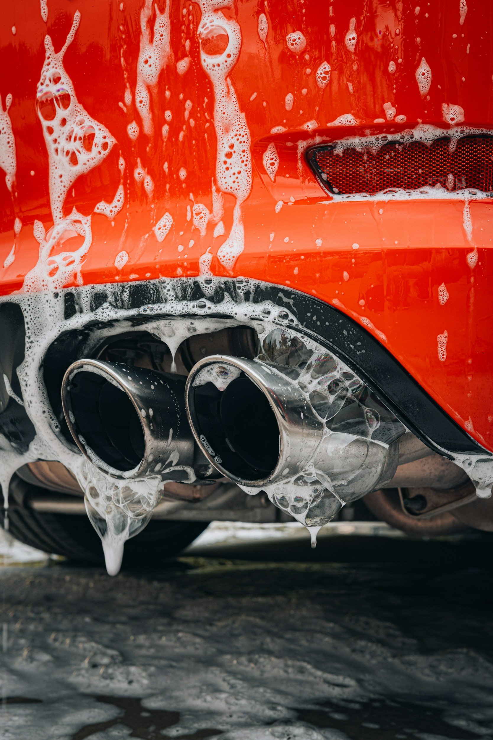 Close-up of the rear of an orange sports car with two exhaust pipes covered in soap suds.