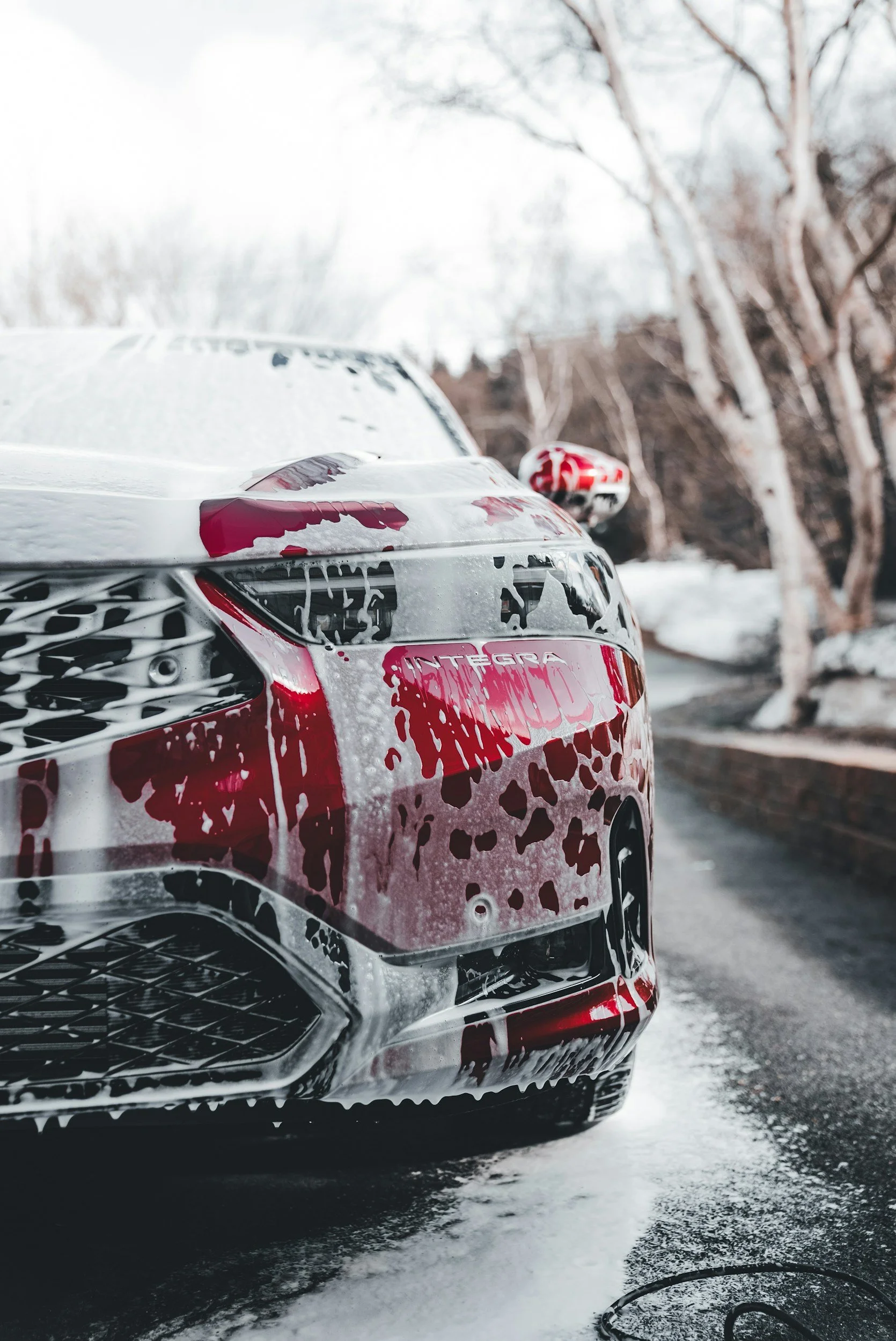 Close-up of a red and white Hyundai Elantra car being washed with soap, on a wet driveway with trees in the background.