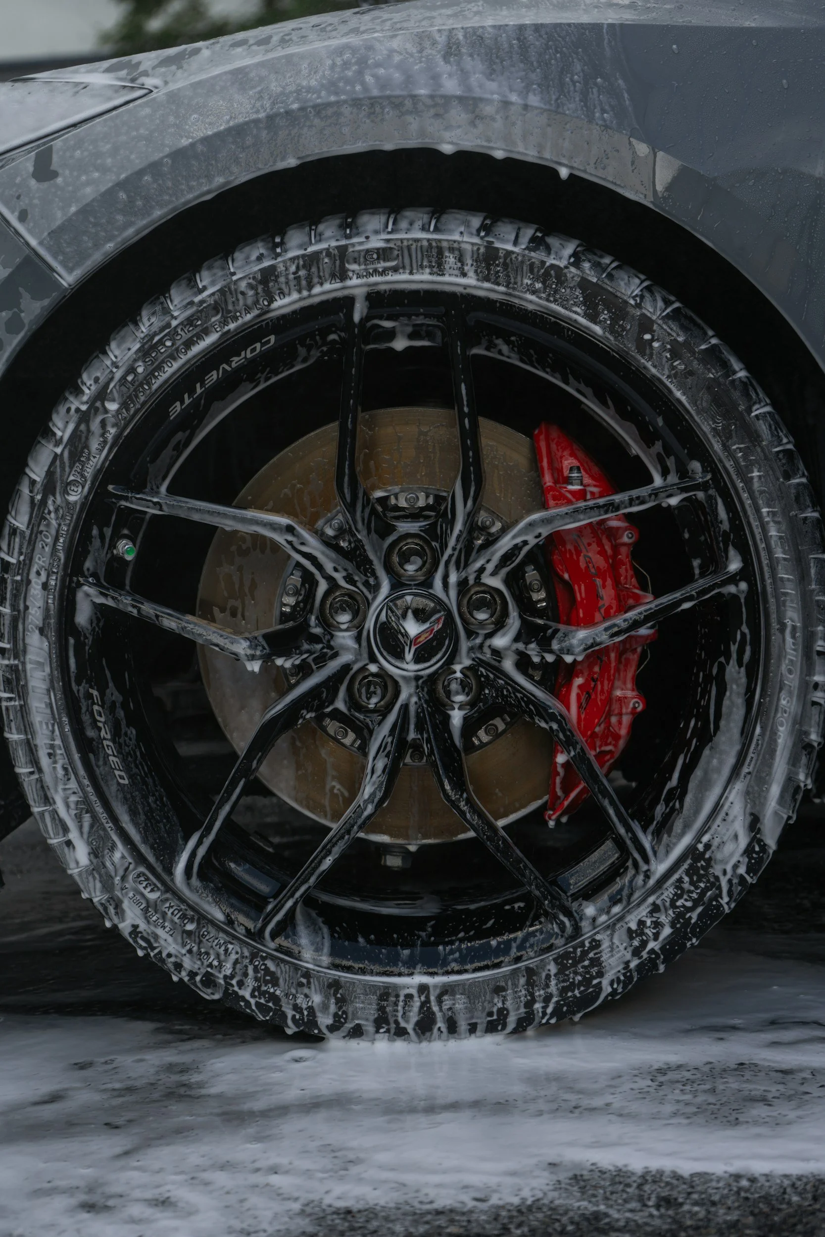 Close-up of a low-profile car tire with foam wash, black alloy wheel, and red brake caliper visible through the wheel spokes, on a wet surface.