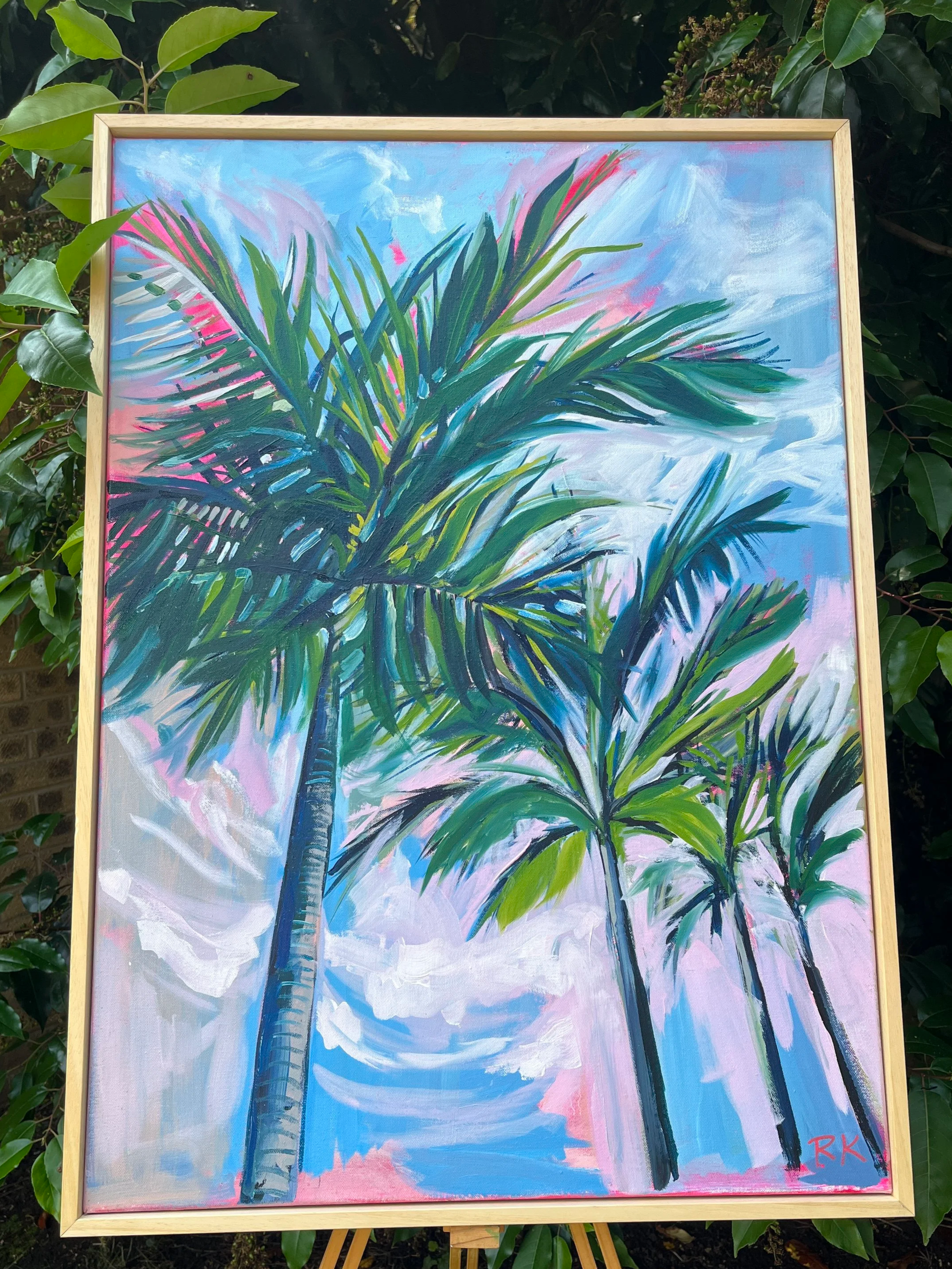 A colorful painting of palm trees against a sky with pink, blue, and white clouds, signed with the initials 'RK' in red in the bottom right corner.
