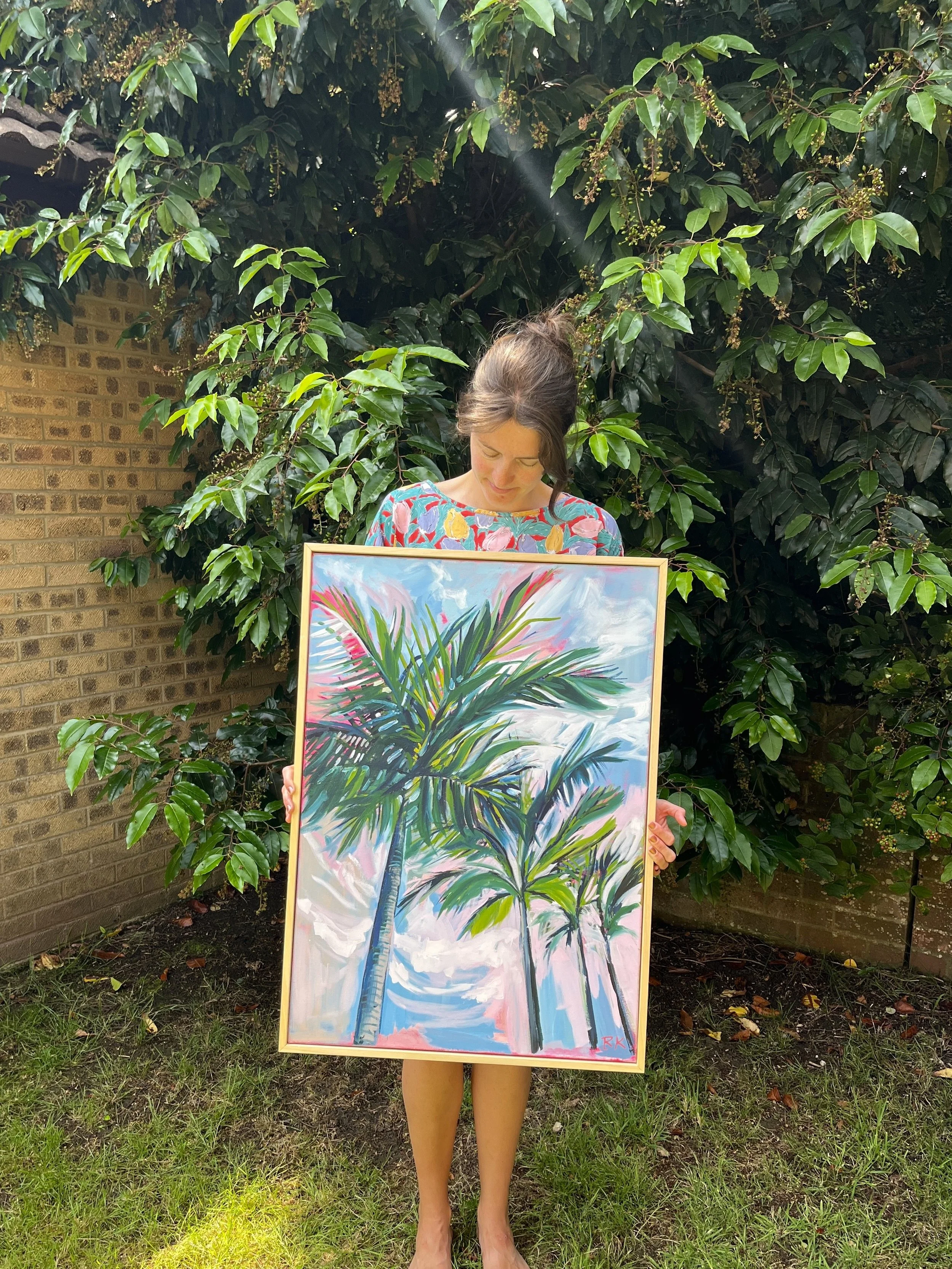 A person standing outdoors on grass, holding a large painting of palm trees against a sky with clouds. The person is wearing a colorful floral dress and has their head bowed, with their face mostly obscured by the top of the painting. There are green