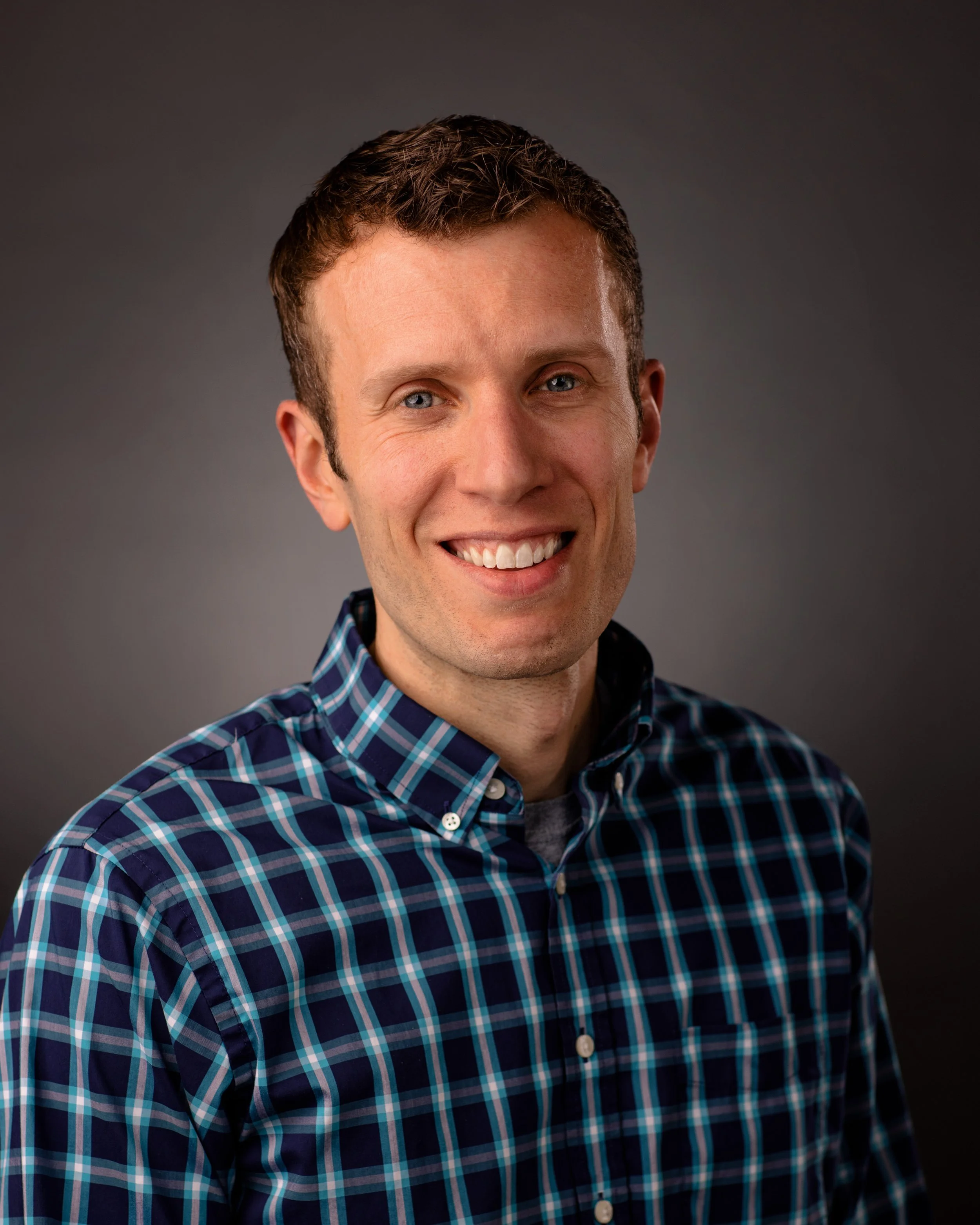 Close-up portrait of a smiling man with short brown hair, blue eyes, wearing a blue and green plaid shirt, against a dark gray background.