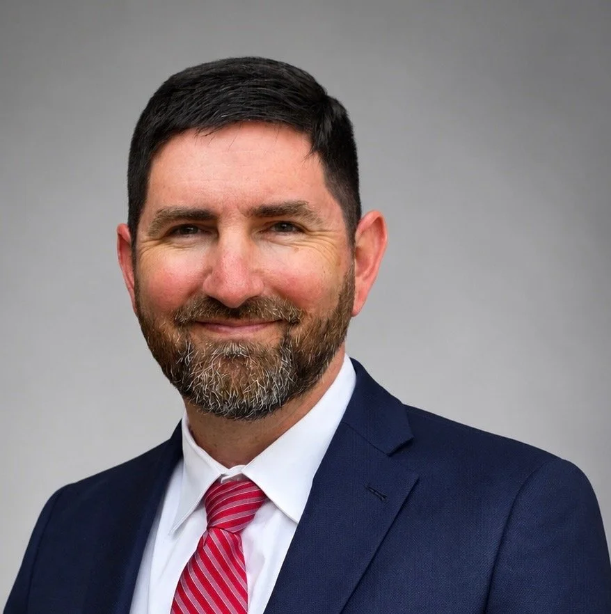 A man with dark hair and a beard is wearing a navy suit, white shirt, and a red striped tie, smiling against a plain gray background.