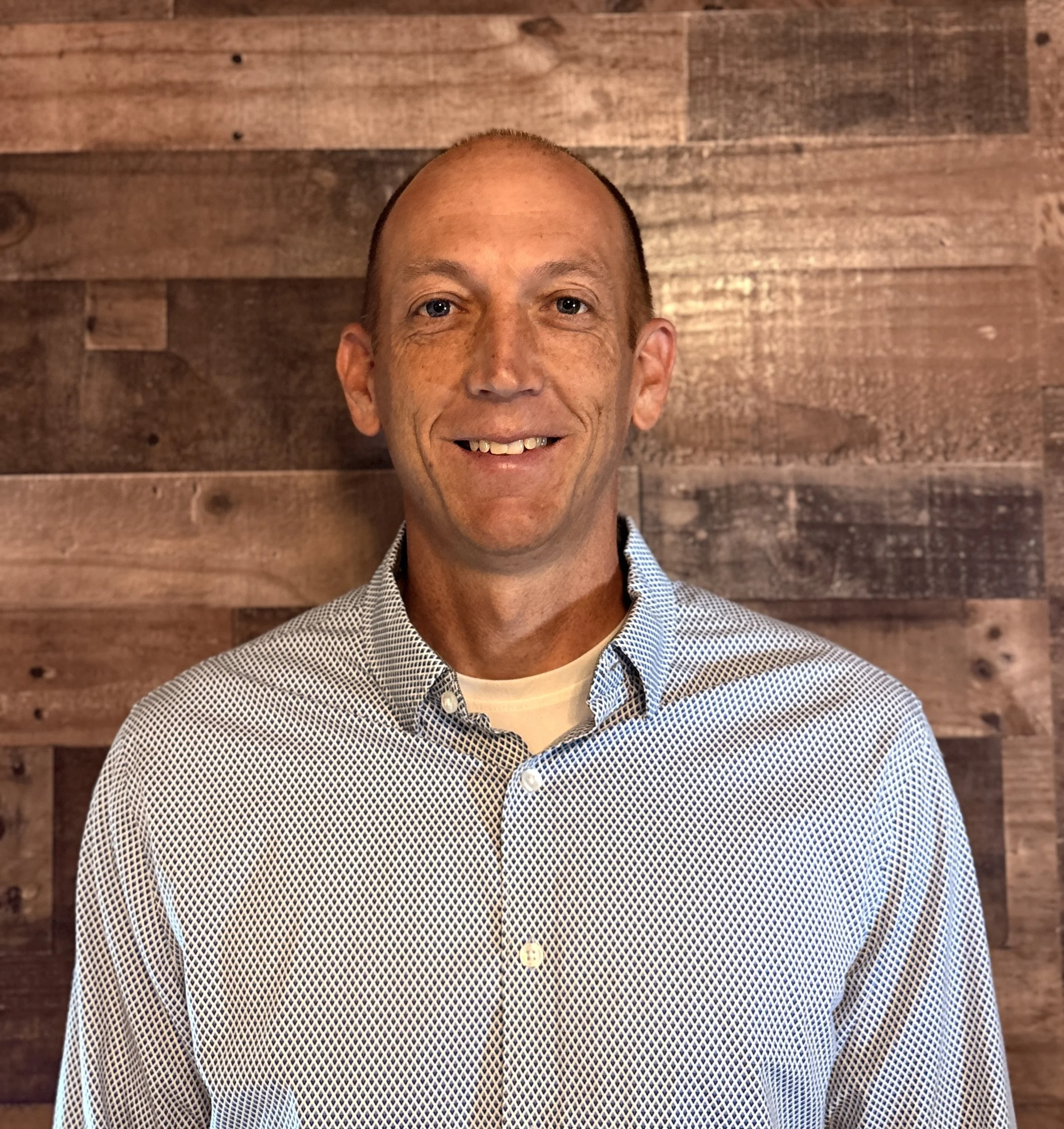 A man with light skin, a smile, and blue eyes, wearing a checkered shirt, is standing in front of a wooden wall.