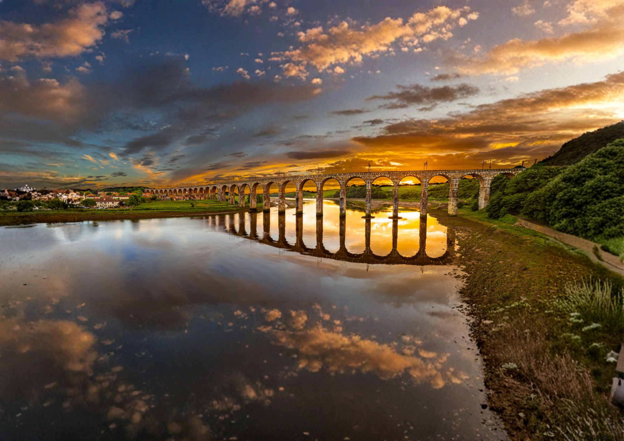 Aerial view of the Royal Border Bridge Viaduct, Berwick Upon Tweed, Northumberland, UK at sunset. Bridge spans a calm river, surrounded by greenery and the town of Berwick Upon Tweed, reflecting the vibrant colours of the sky filled with clouds.