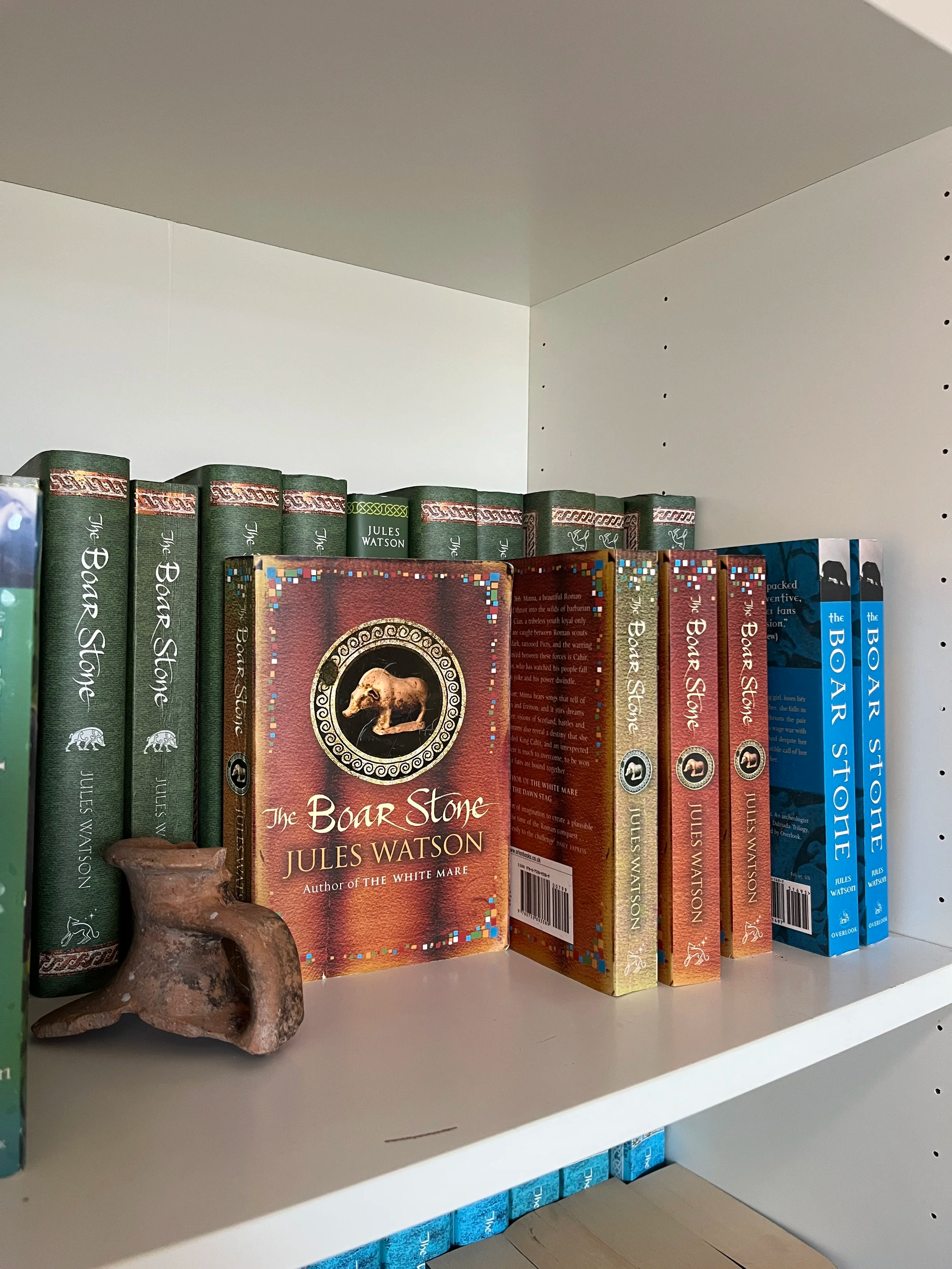 Photo shows a bookshelf containing several books by Jules Watson, including several editions of 'The Boar Stone'. There is a small, brown section of pottery in front of the books.