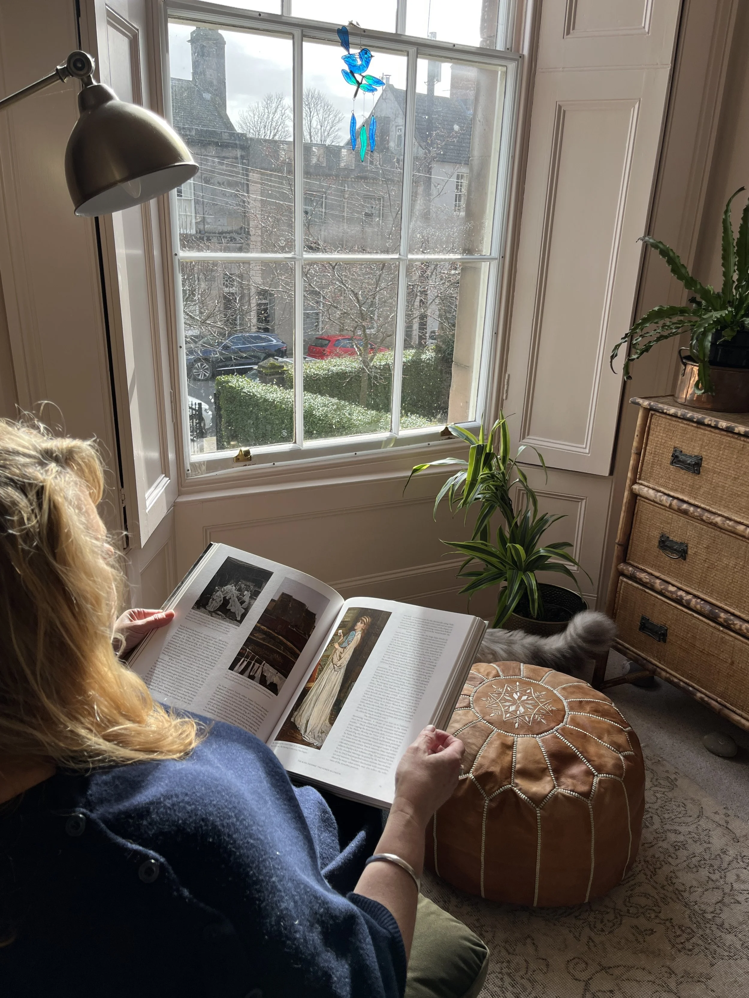 Photo shows author Jules Watson reading in a chair opposite a large window. There is a green plant on a chest of drawers and sunlight coming through the window, with a blue and green stained glass wind chime hanging outside.