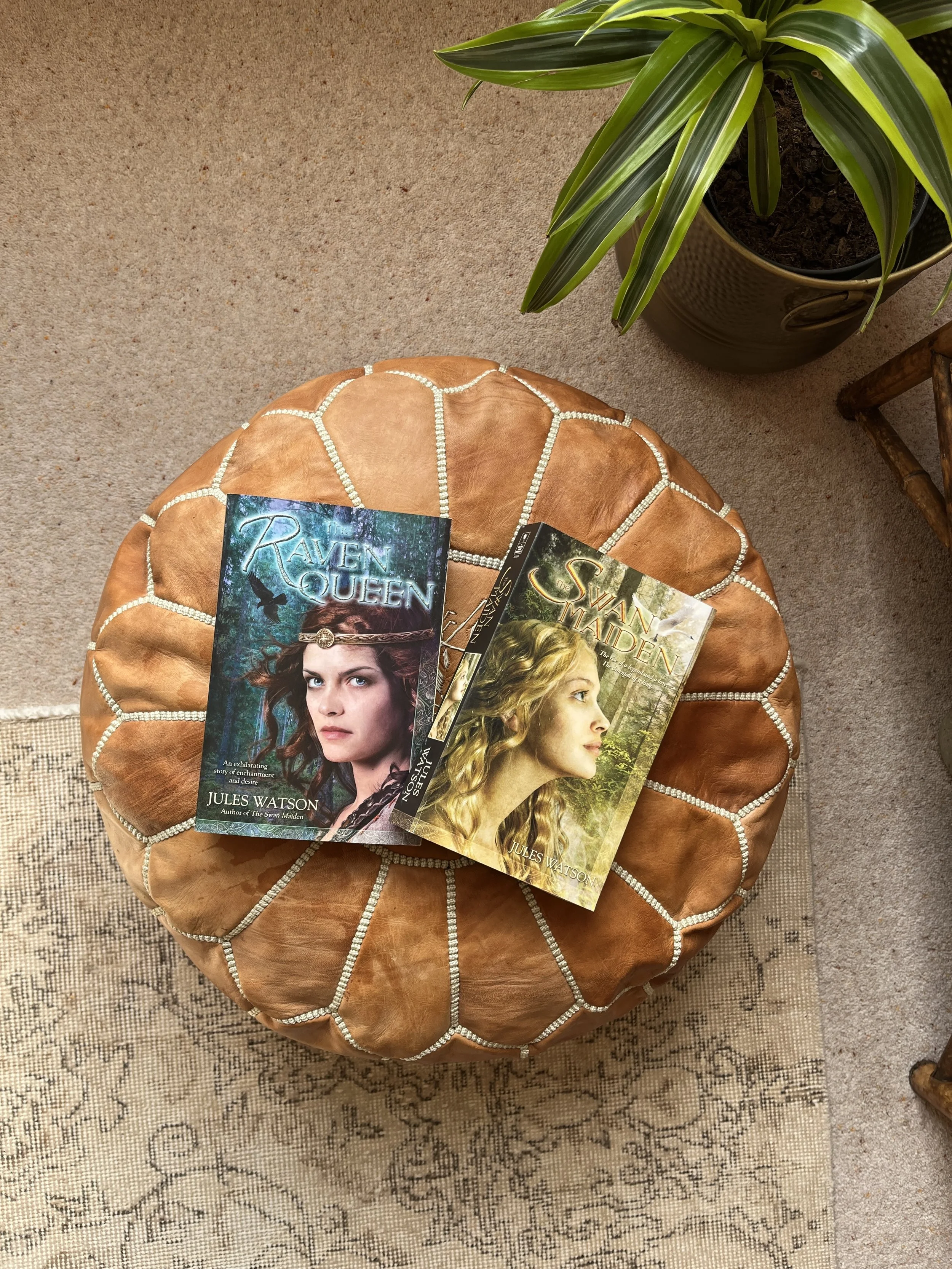 Photo shows two books by Jules Watson, ‘The Raven Queen’ and ‘The Swan Maiden’ placed on a round, padded, tan-colored stool with light coloured stitching. A potted green plant is in the background on a beige carpeted floor.