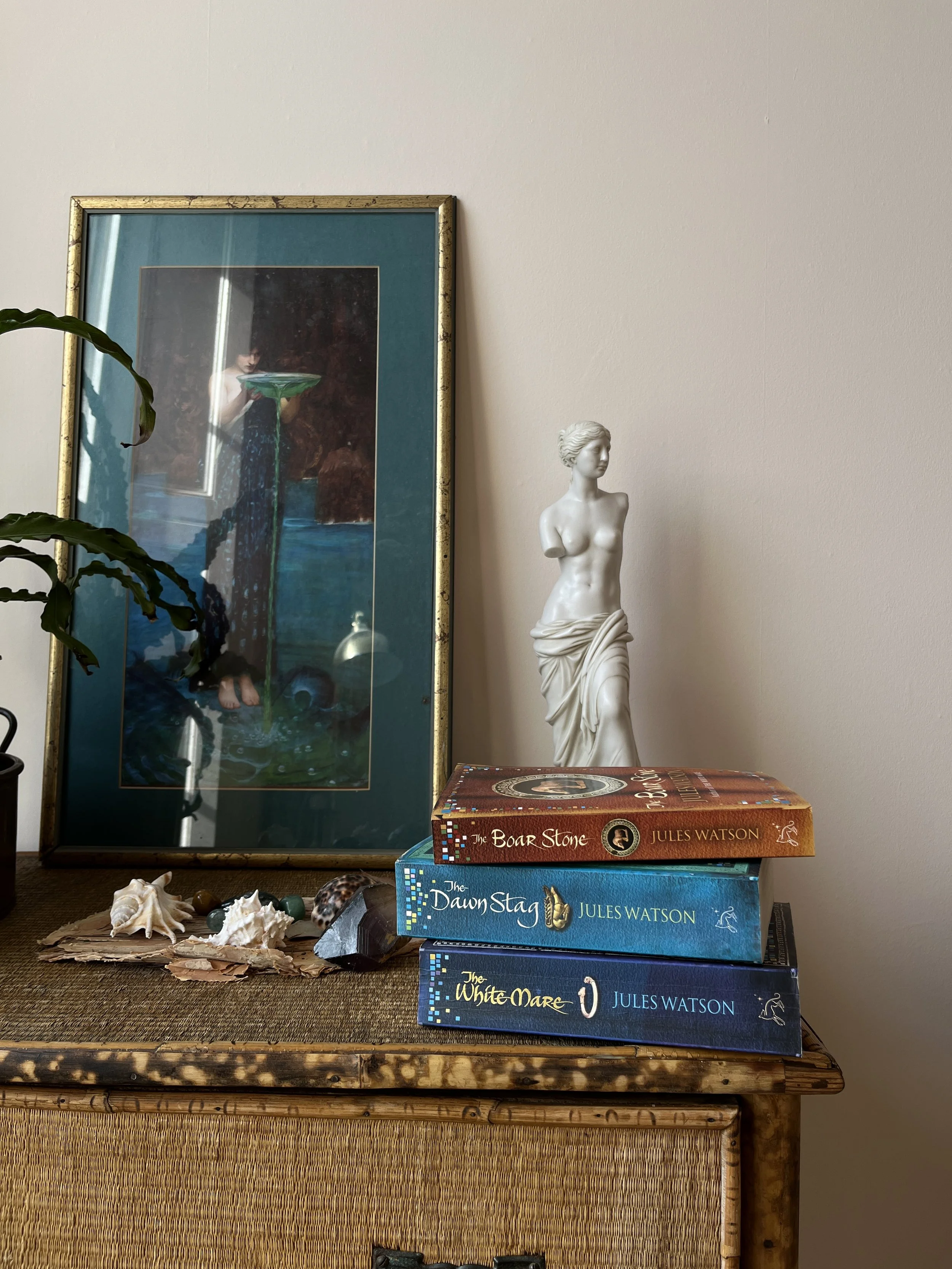 Photo showing chest top with seashells and stones and three books by Jules Watson titled 'The Boar Stone,' 'The Dawn Stag,' and 'The White Mare.' Behind books is ‘Circe Invidiosa’ by John William Waterhouse and a white female bust sculpture.