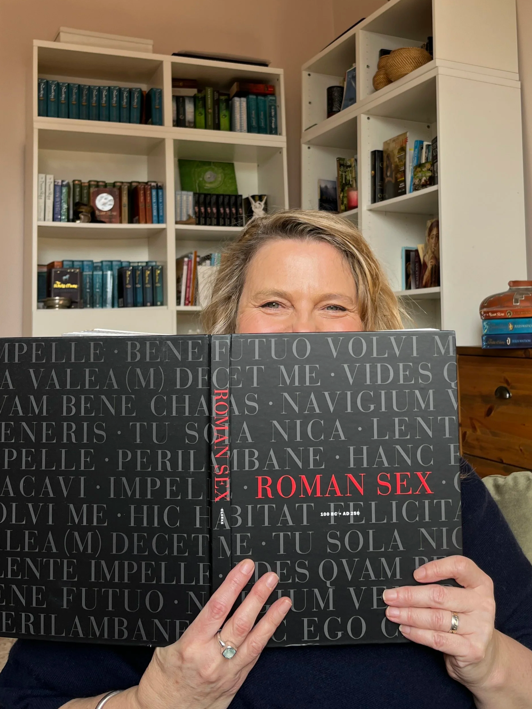 Photo of author Jules Watson holding a large book titled 'Roman Sex' in front of her face, with part of her face visible above the book. She has blonde, wavy hair. There are bookshelves in the background filled with various books.
