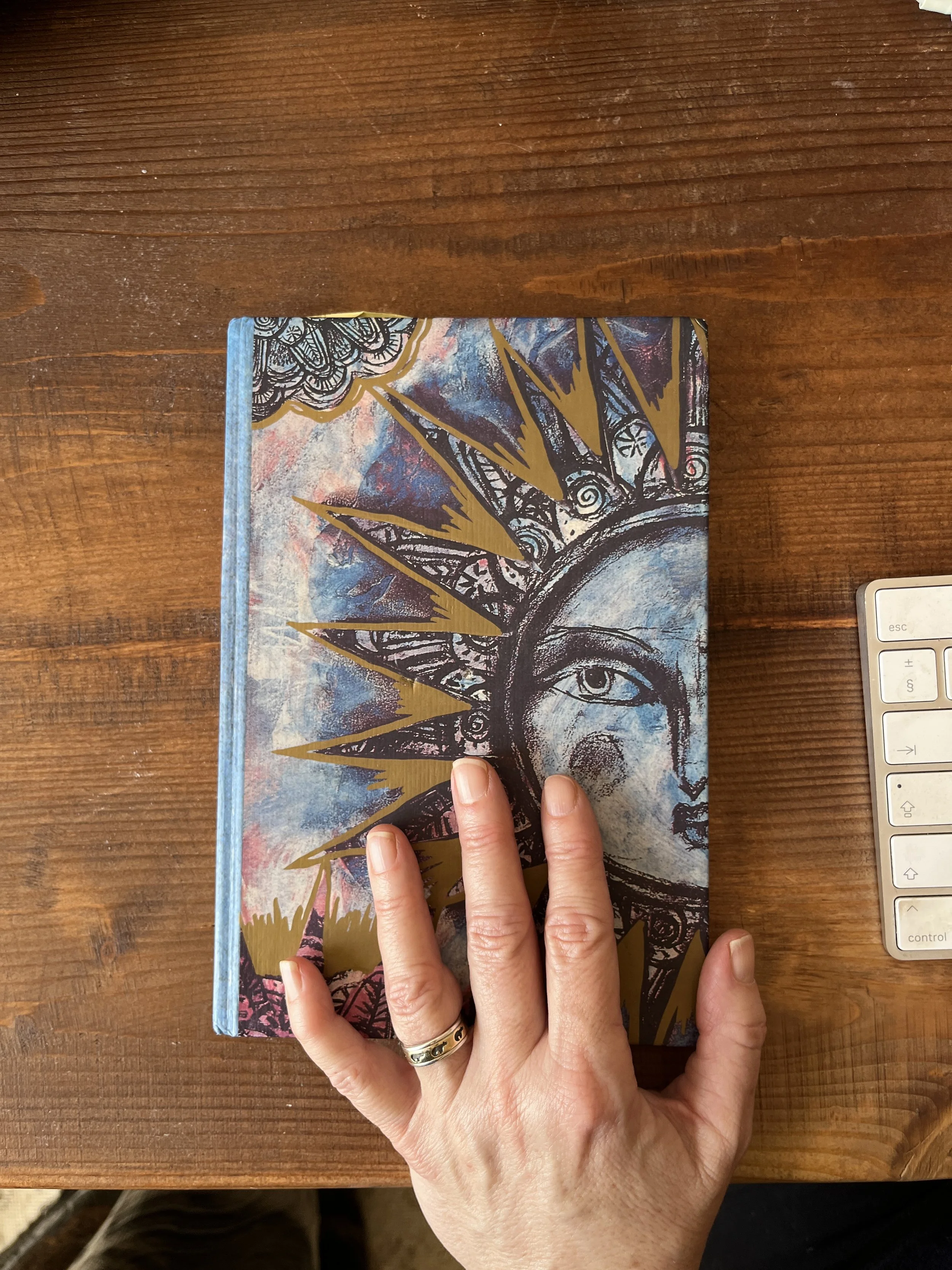 Photo shows the hand of author Jules Watson which is placed on a notebook next to a keyboard. The cover of the notebook features a stylized face with a sun/ crown motif and intricate patterns.