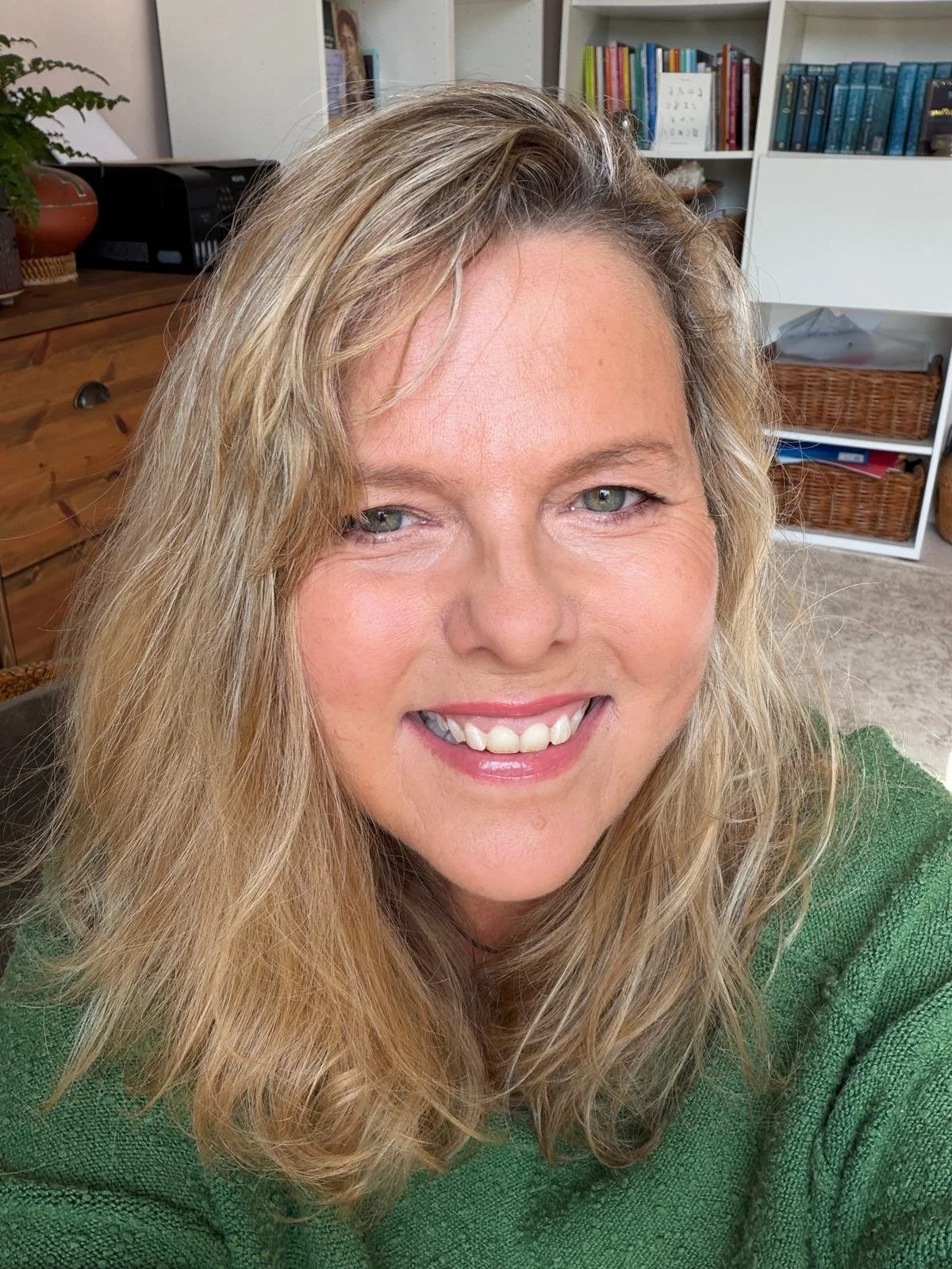 Close-up photo of author Jules Watson smiling. Jules Watson has long blonde hair and is wearing a green top. In the background are bookshelves which contain some of her books.