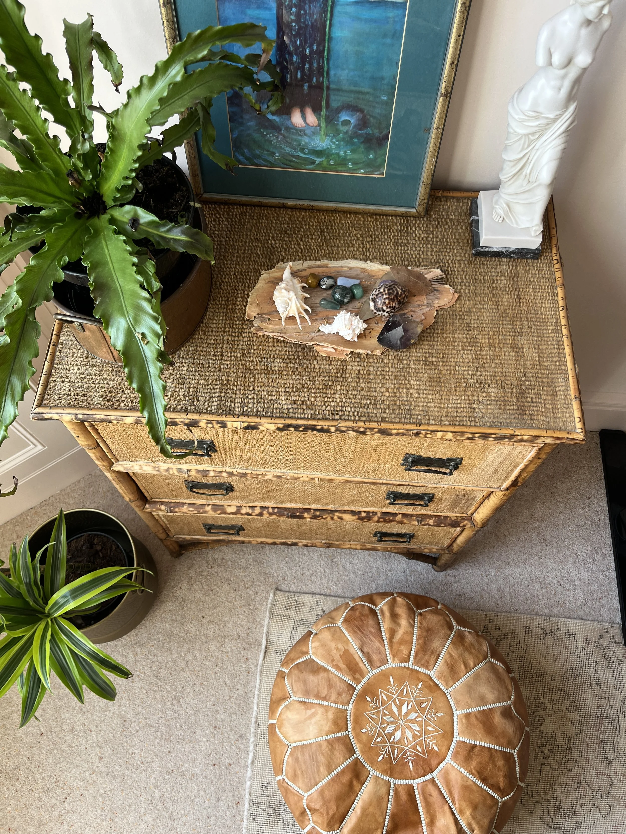 Photo shows a wicker chest of drawers from above with various seashells and stones arranged on a wooden tray on the top of it. A tall white sculpture of a woman's torso stands to the right, and there are green indoor plants nearby.