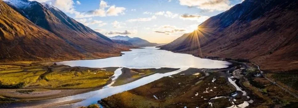 Photo of Loch Etive in the Scottish Highlands. A river flows between mountain valleys with low vegetation, as the sun sets behind the mountains casting a warm glow.