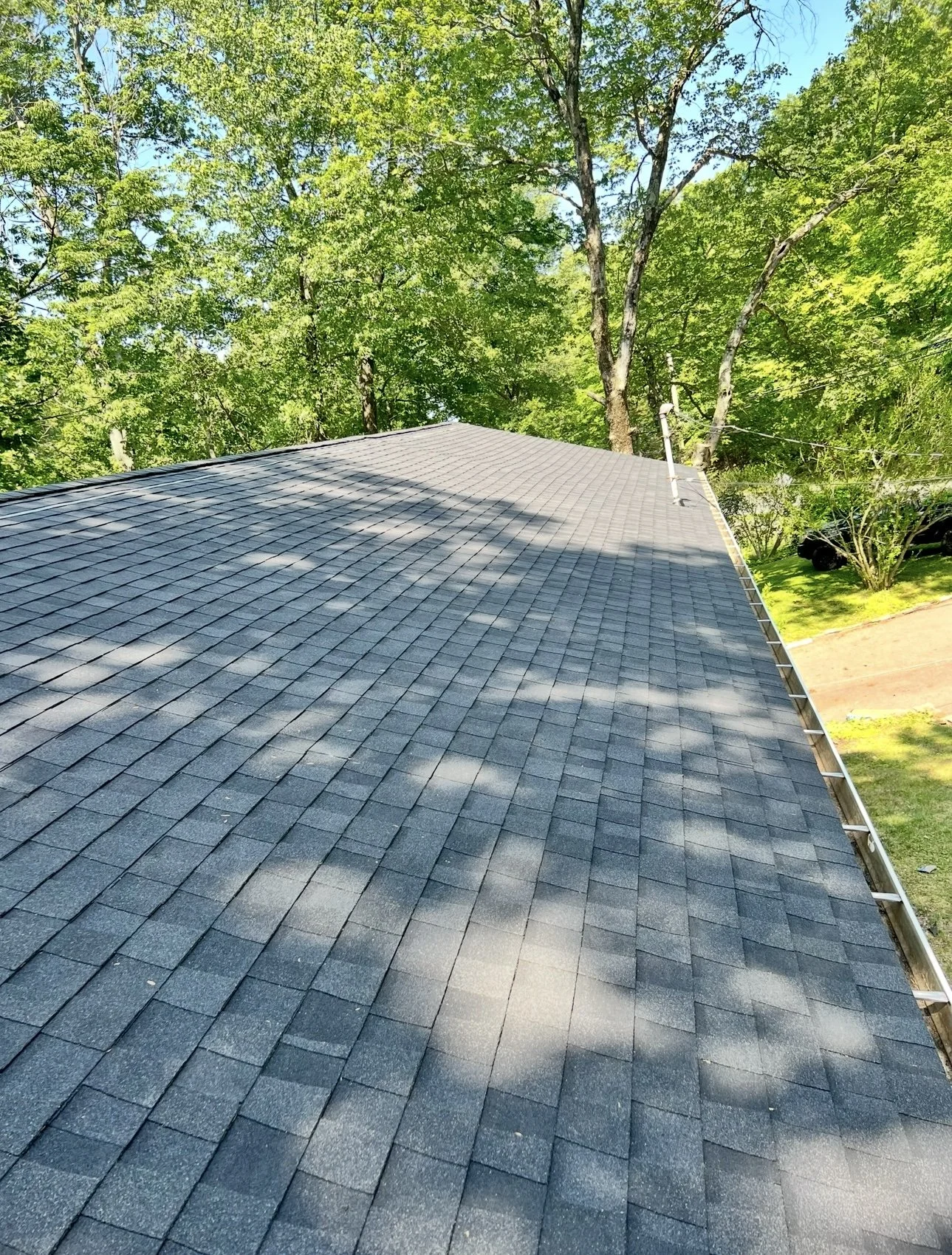 View of a house roof with gray asphalt shingles, surrounded by green trees and a bright blue sky.