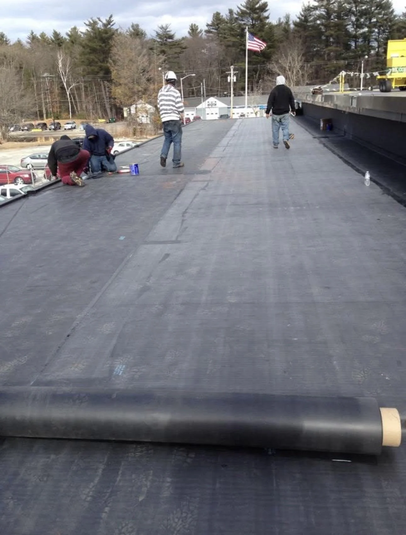 Workers installing roofing on a building's flat roof, with a water bottle and tools visible, and an American flag in the background.