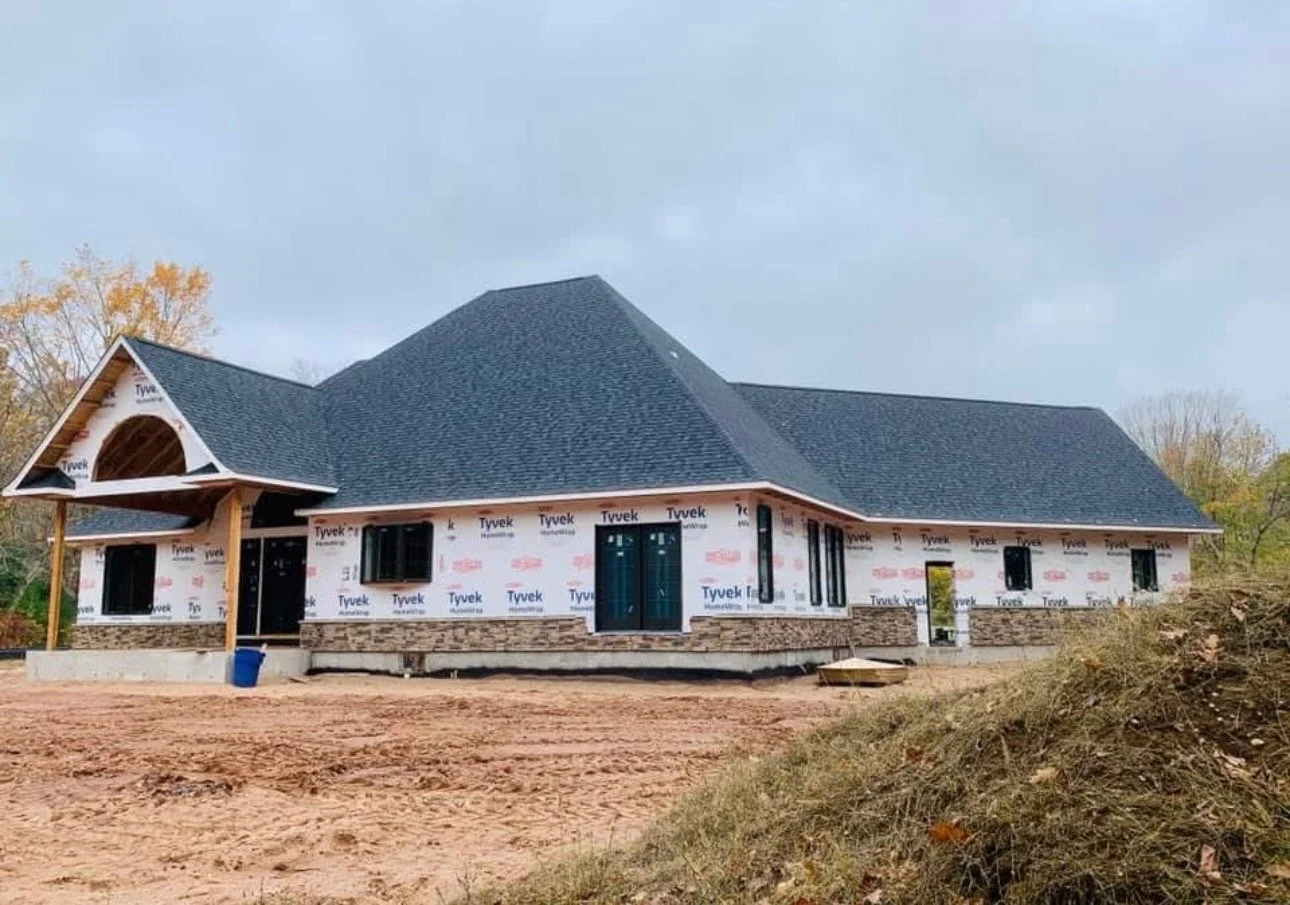 A house under construction with black shingles, partly wrapped in Tyvek. The house has a stone foundation and multiple windows, and the surrounding area appears to be a dirt lot with no grass yet.