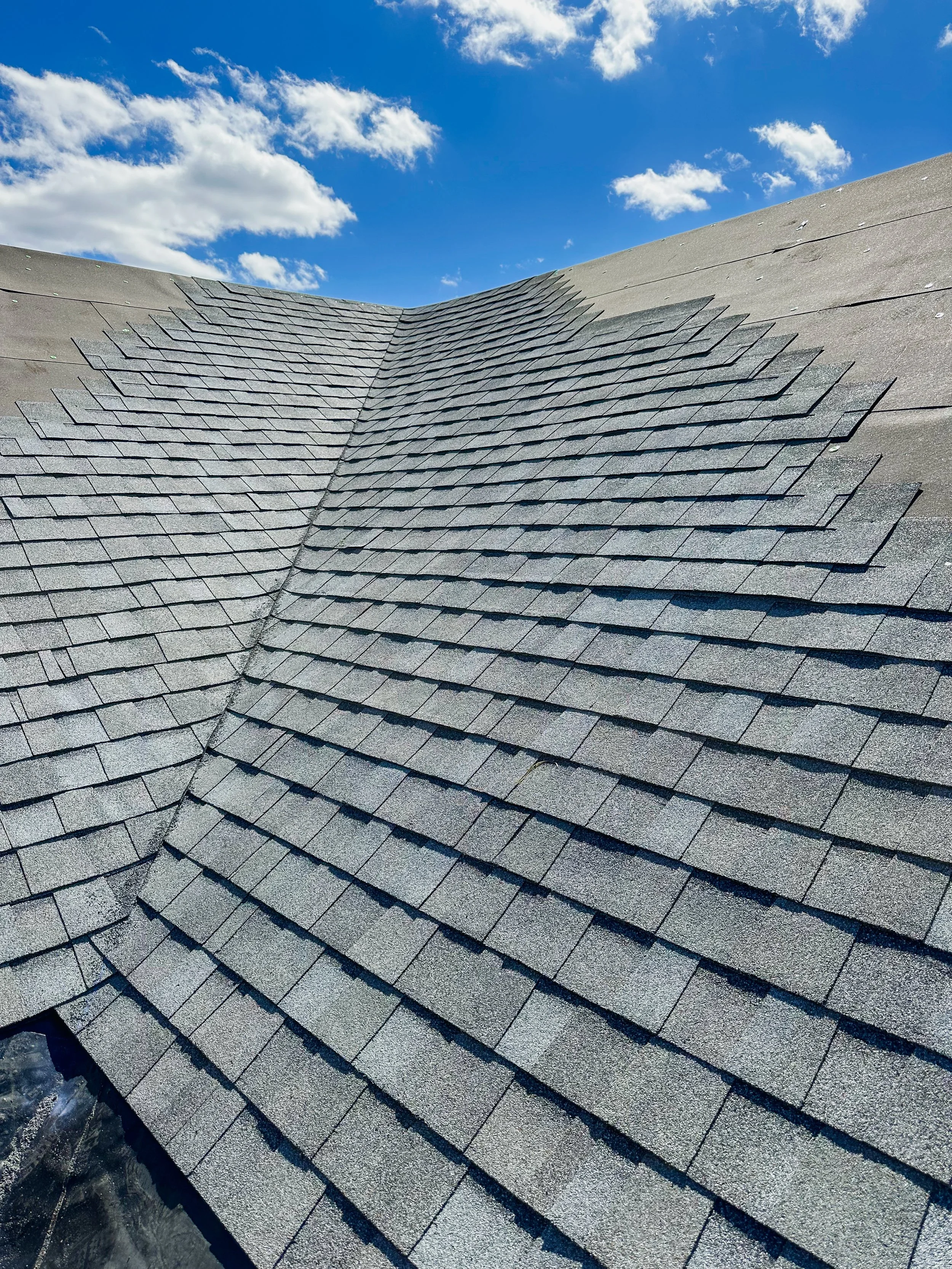 Close-up of a gabled roof with gray asphalt shingles and a small section of flashing at the bottom corner, under a blue sky with white clouds.