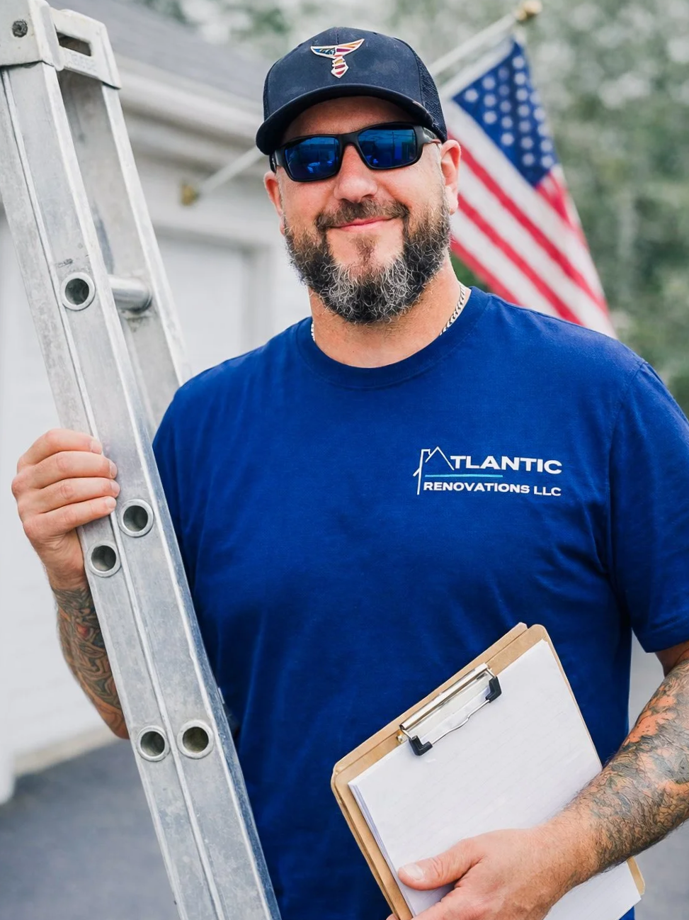 A man smiling outdoors, wearing sunglasses, a black cap, and a blue t-shirt with a construction company logo. He's holding a ladder and a clipboard, with an American flag in the background.