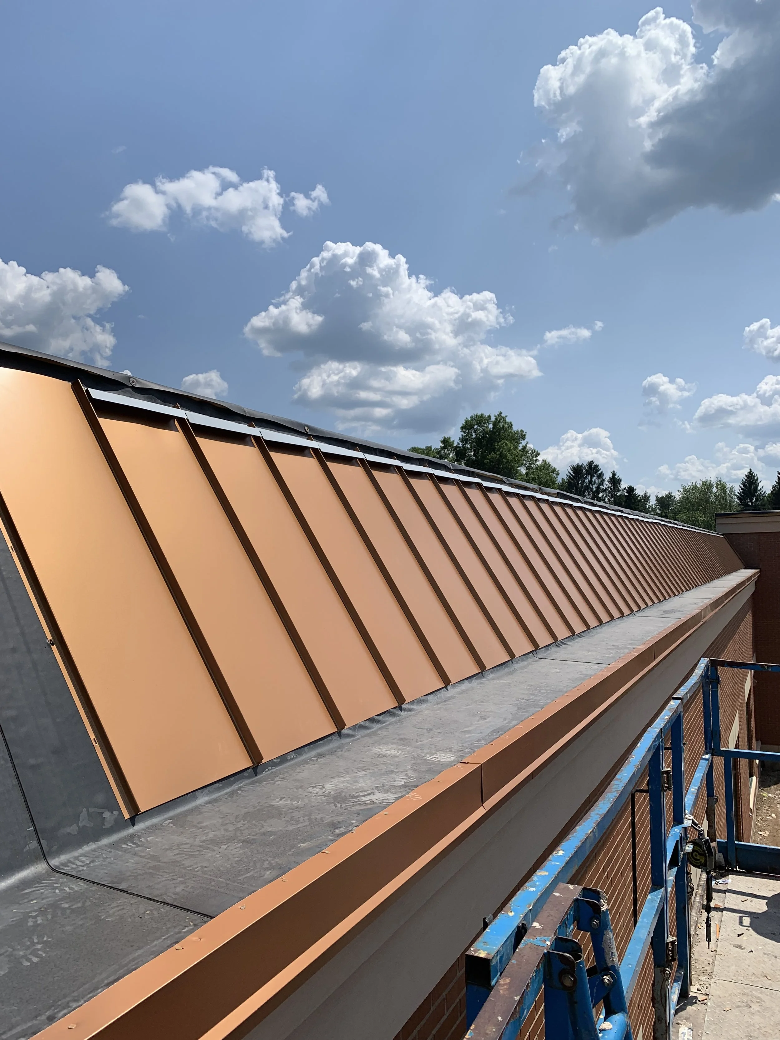 Copper roof panels installed on a building under a partly cloudy sky, with some trees visible in the background.