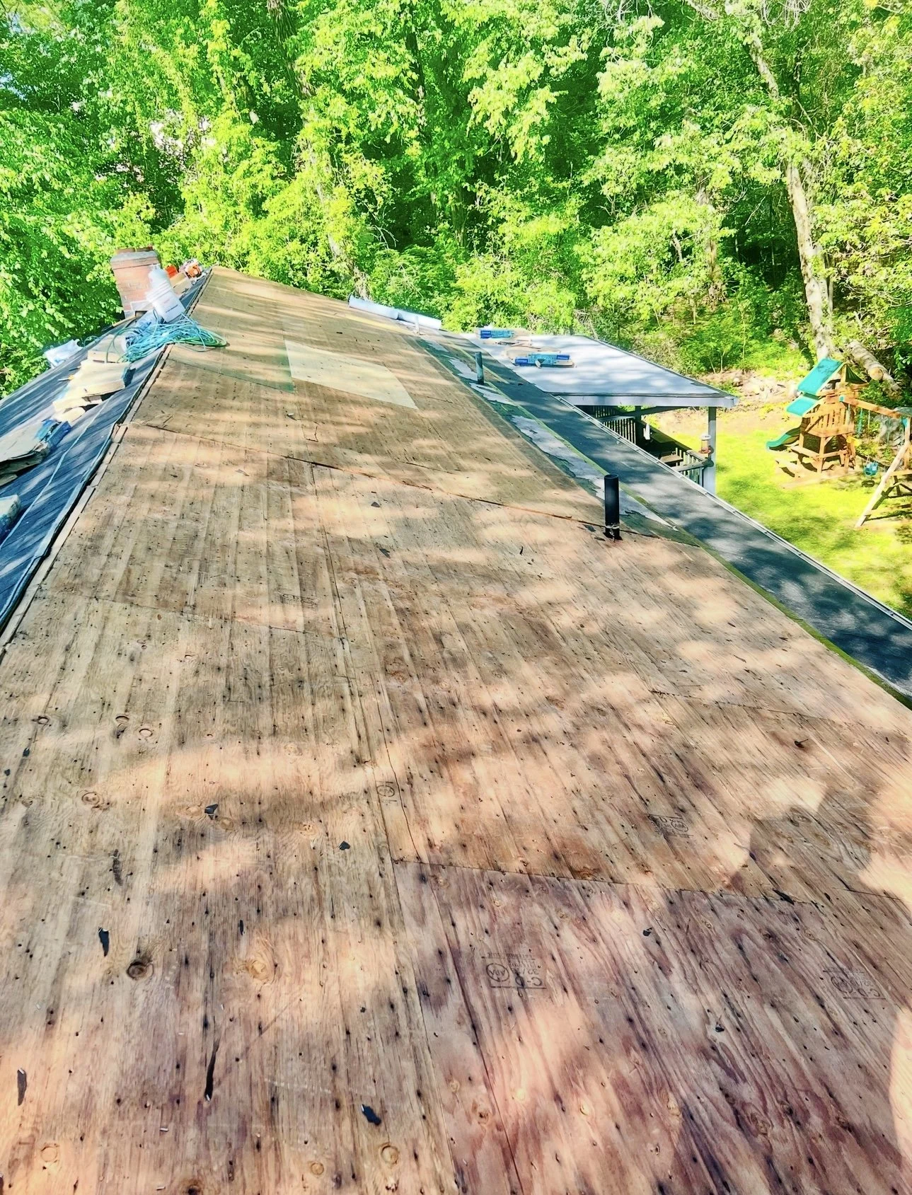 Roof under renovation with plywood sheathing, surrounding trees and a backyard with a slide and a playhouse.